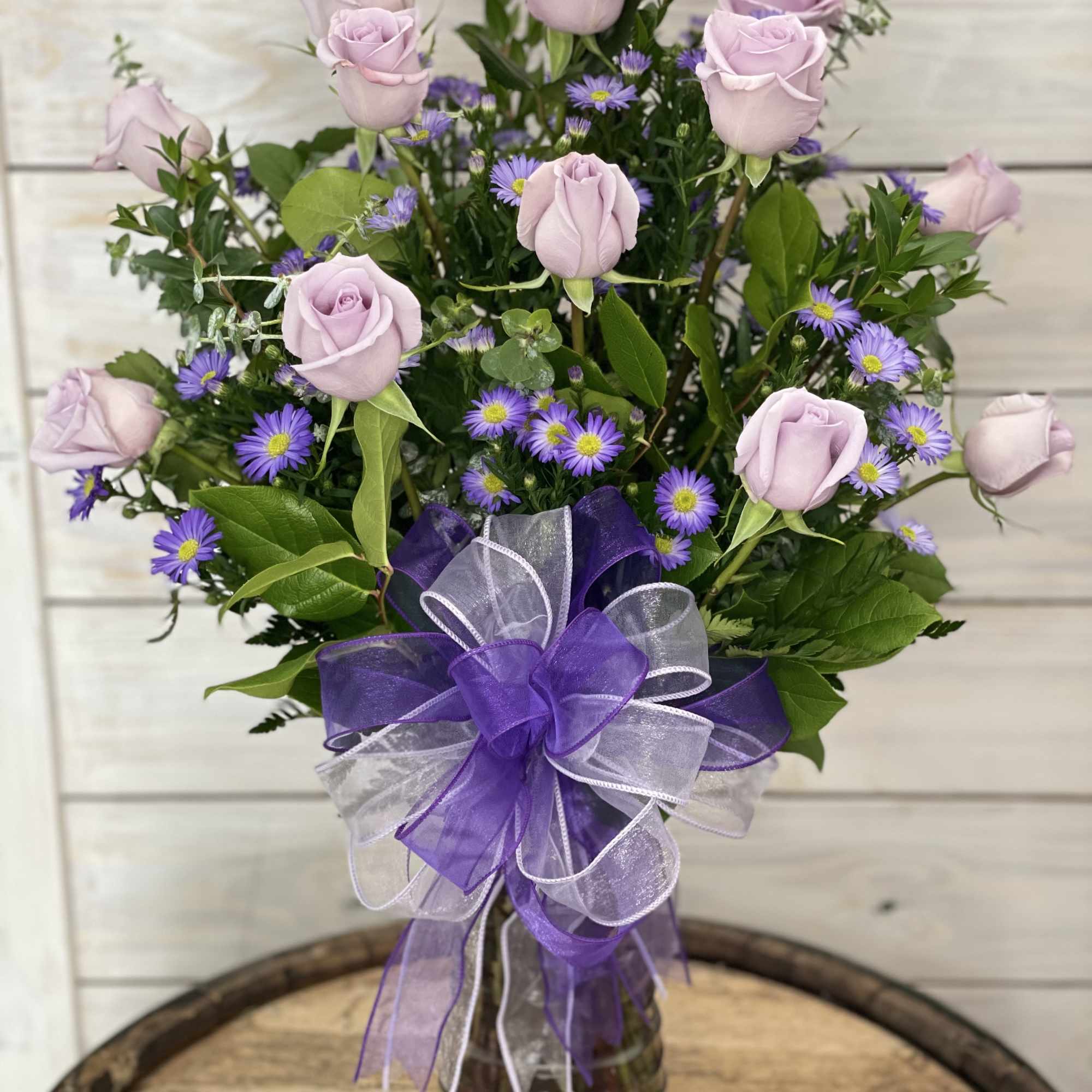 Lavender roses and purple daisies in a glass vase with a purple ribbon
