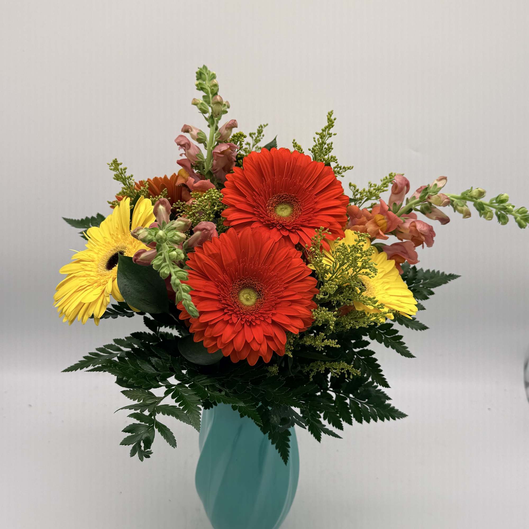 Arrangement of red and yellow gerbera daisies with pink snapdragons in a teal vase