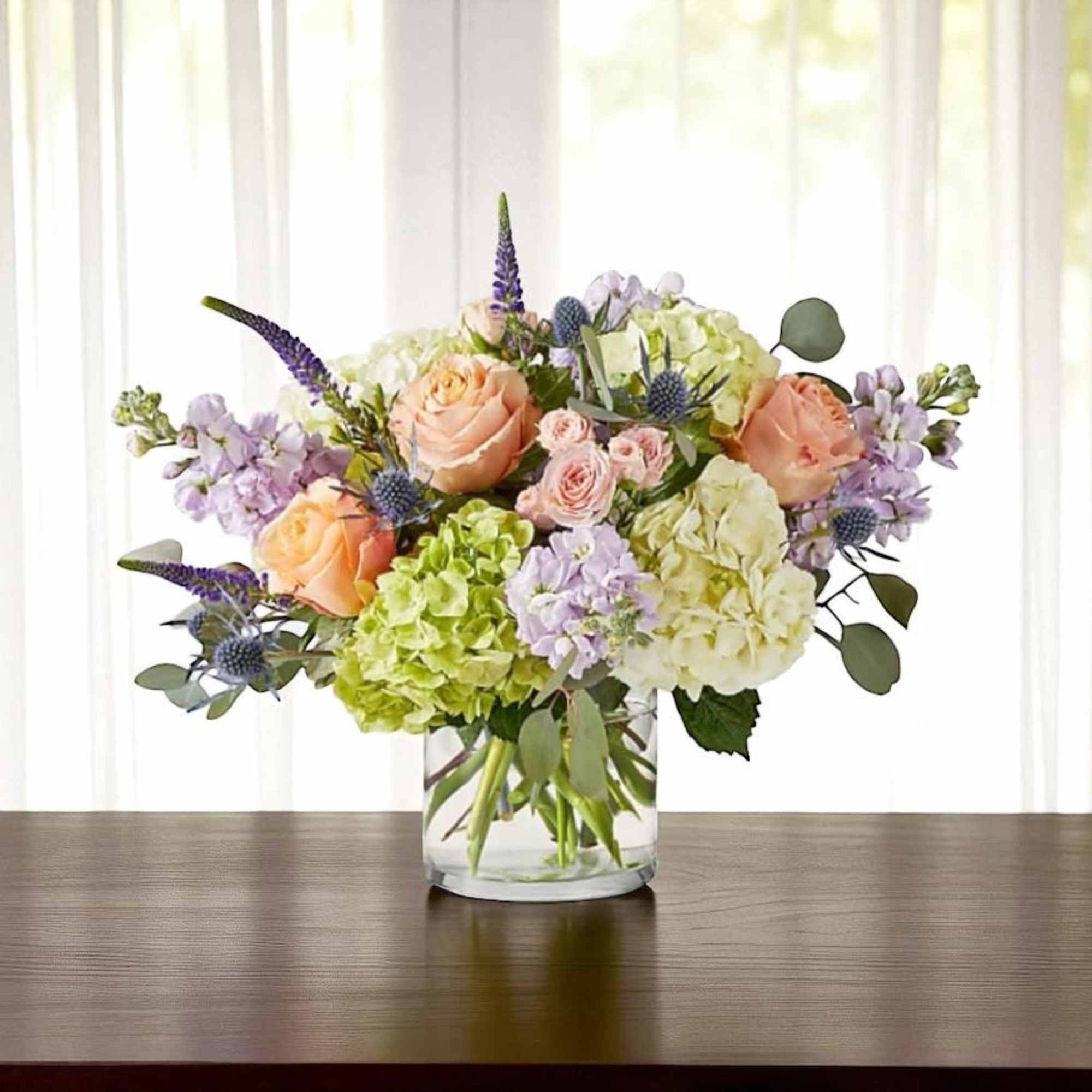 Peach roses, green and white hydrangeas, and lavender blooms in a clear glass cylinder vase