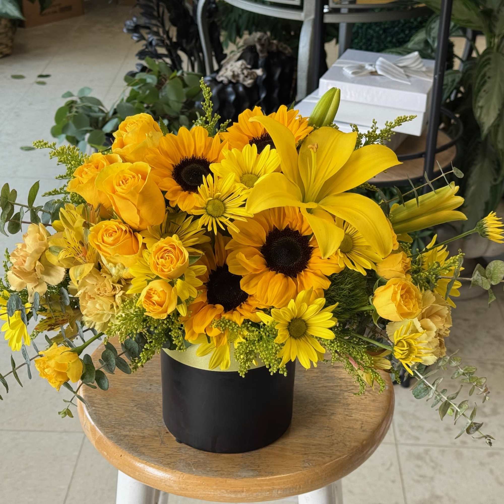 Bright yellow arrangement of sunflowers, lilies, roses, and daisies in a black cylinder vase