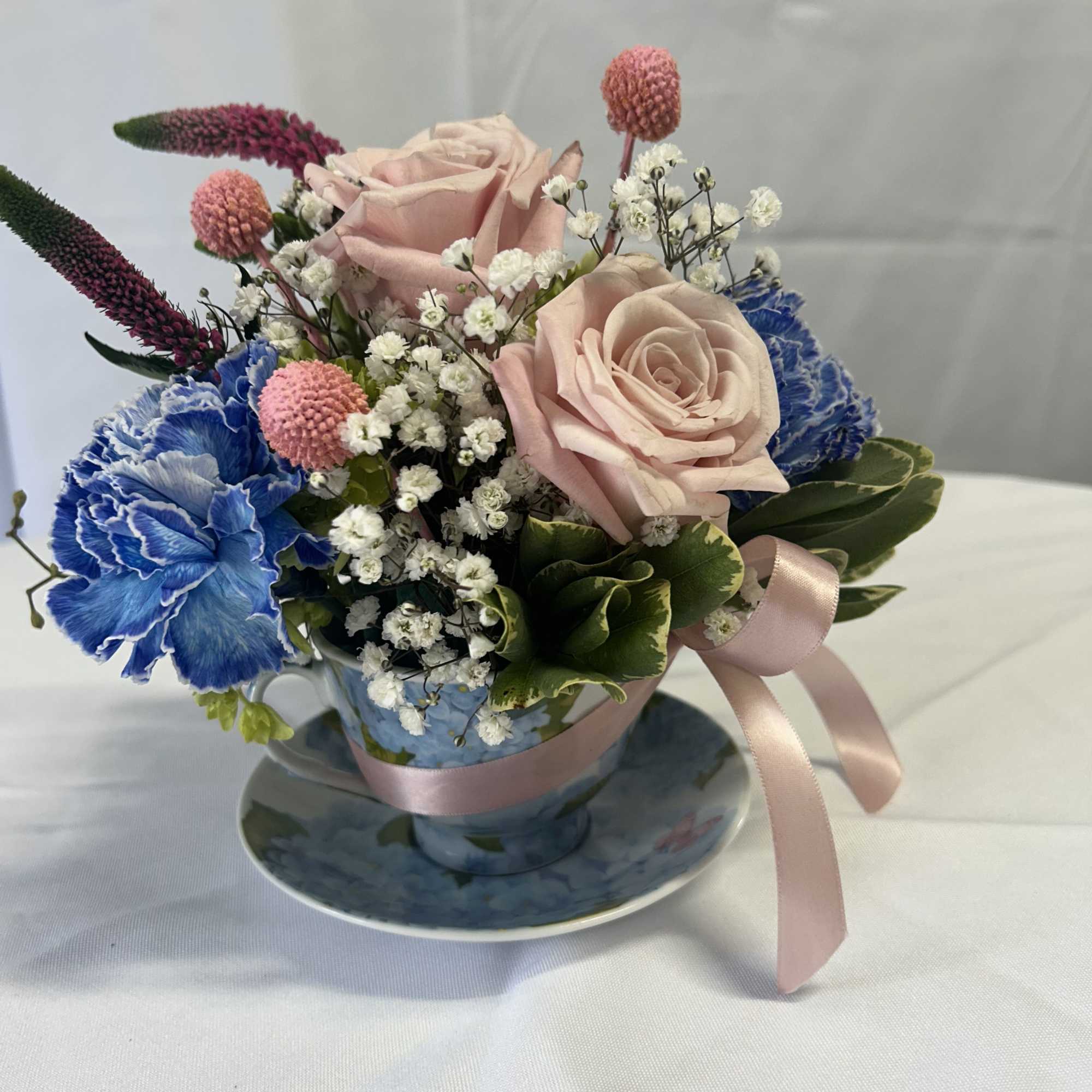 Pink roses and blue carnations arranged in a teacup with a ribbon