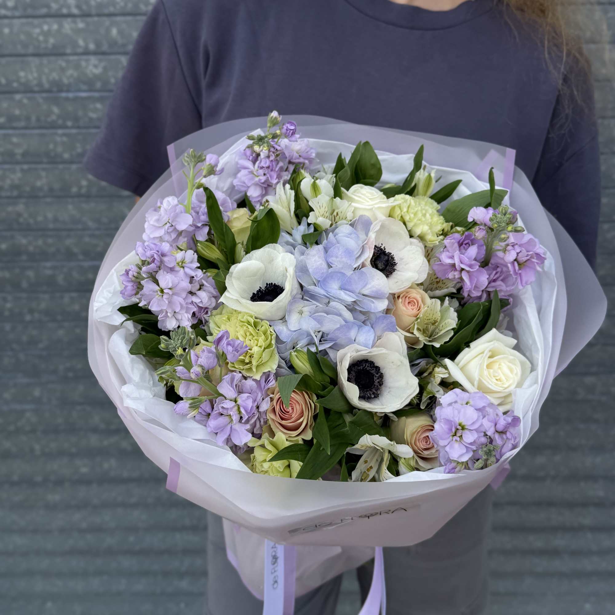Hand-tied bouquet of white, peach, and lavender flowers wrapped in pale pink paper, held by a person in a gray shirt.