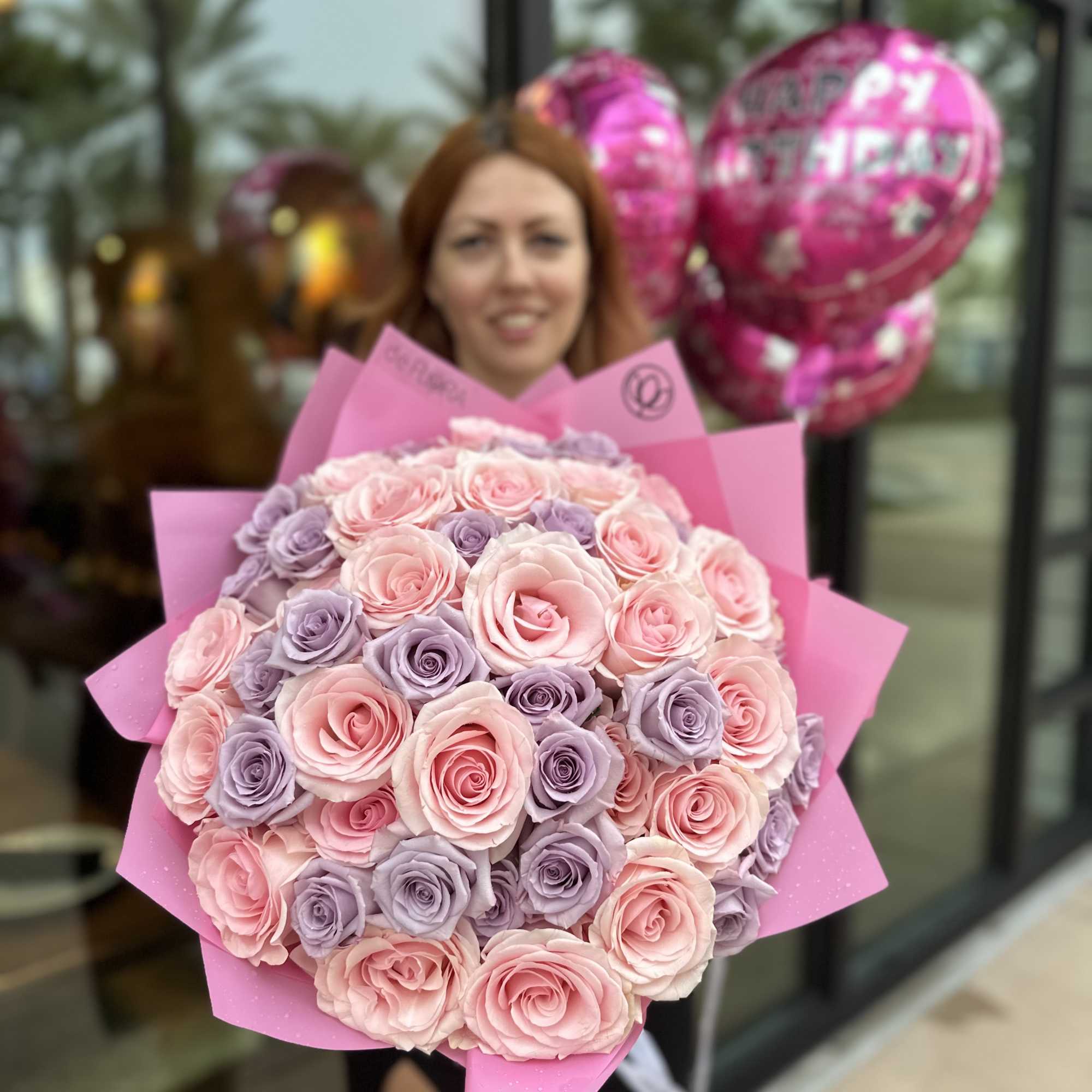 Large bouquet of pink and lavender roses wrapped in pink paper, held in front of birthday balloons