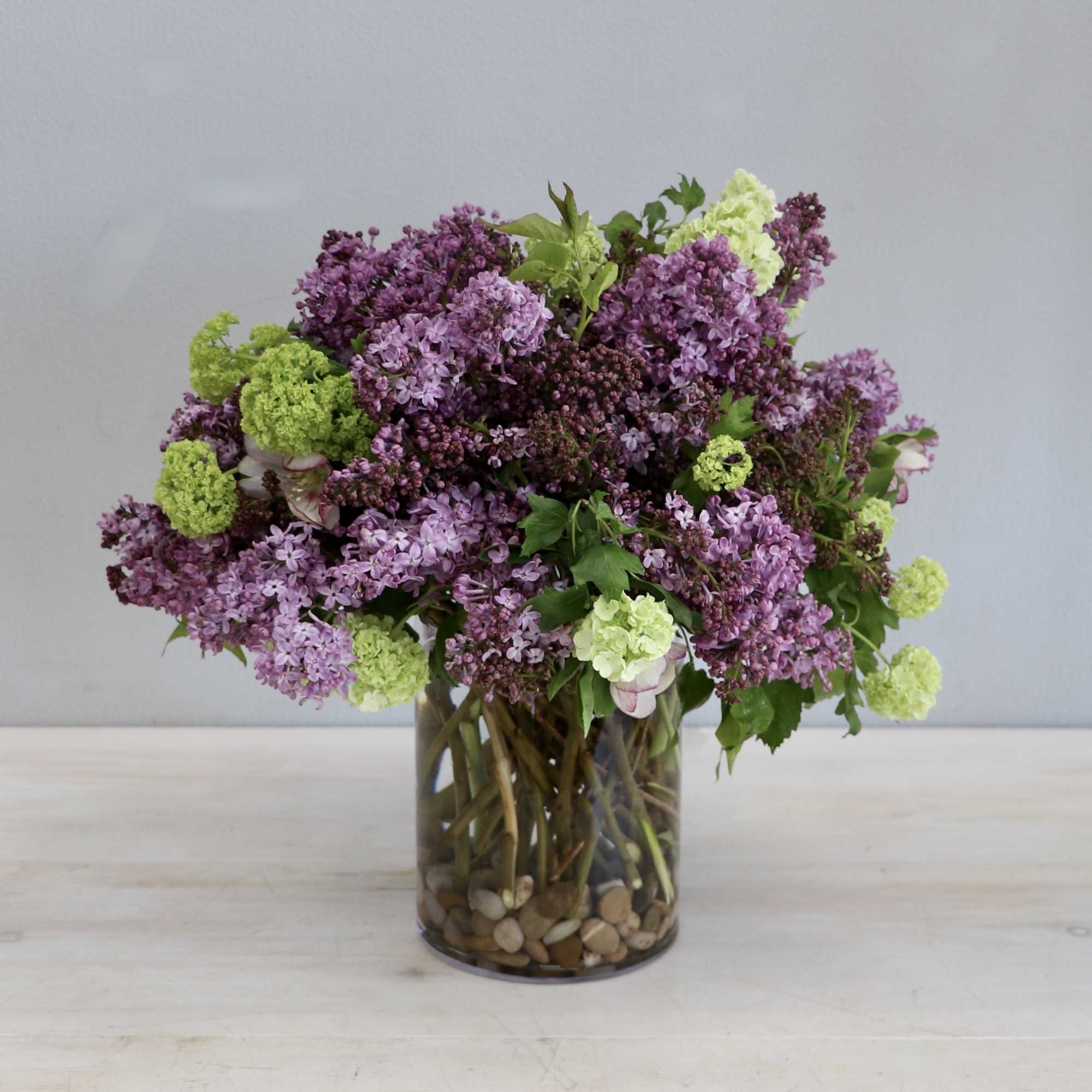 Purple lilacs and green blooms arranged in a glass vase with pebbles