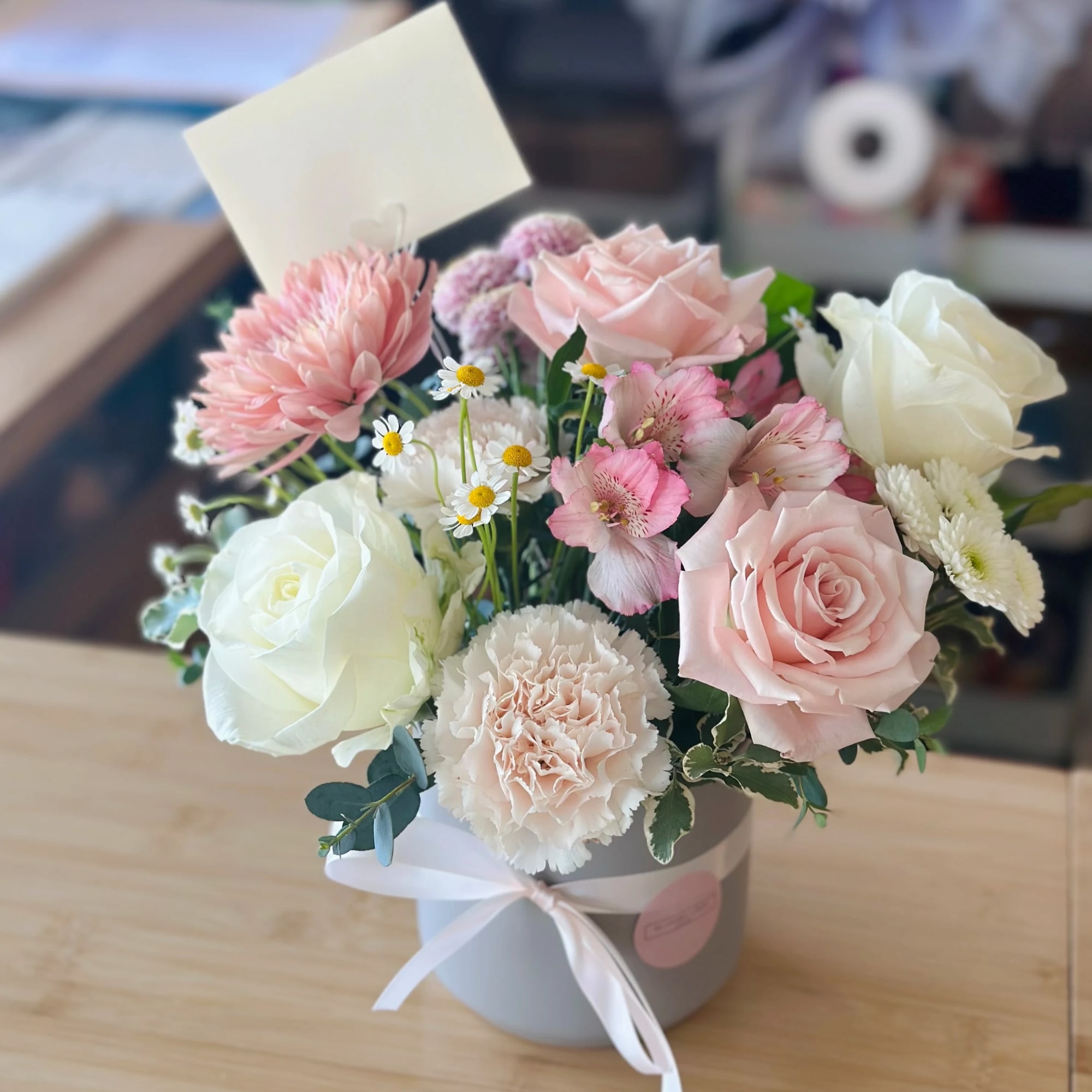Pastel arrangement of pink and white roses, carnations, and daisies in a white hatbox with ribbon and card.