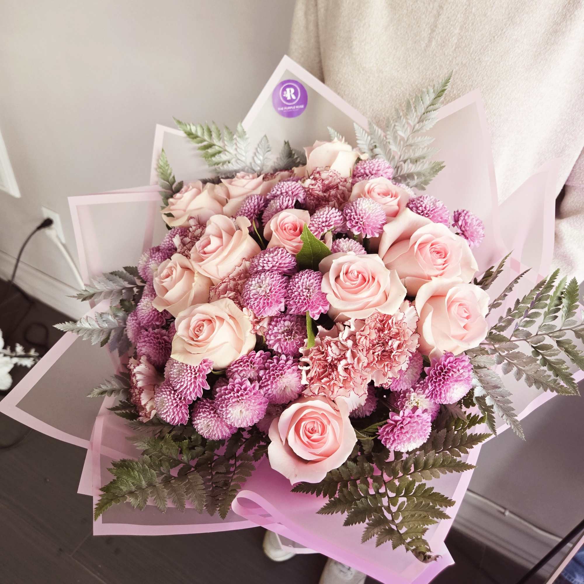 Hand-tied bouquet of light pink roses, pink carnations, and pompom blooms wrapped in pink paper.