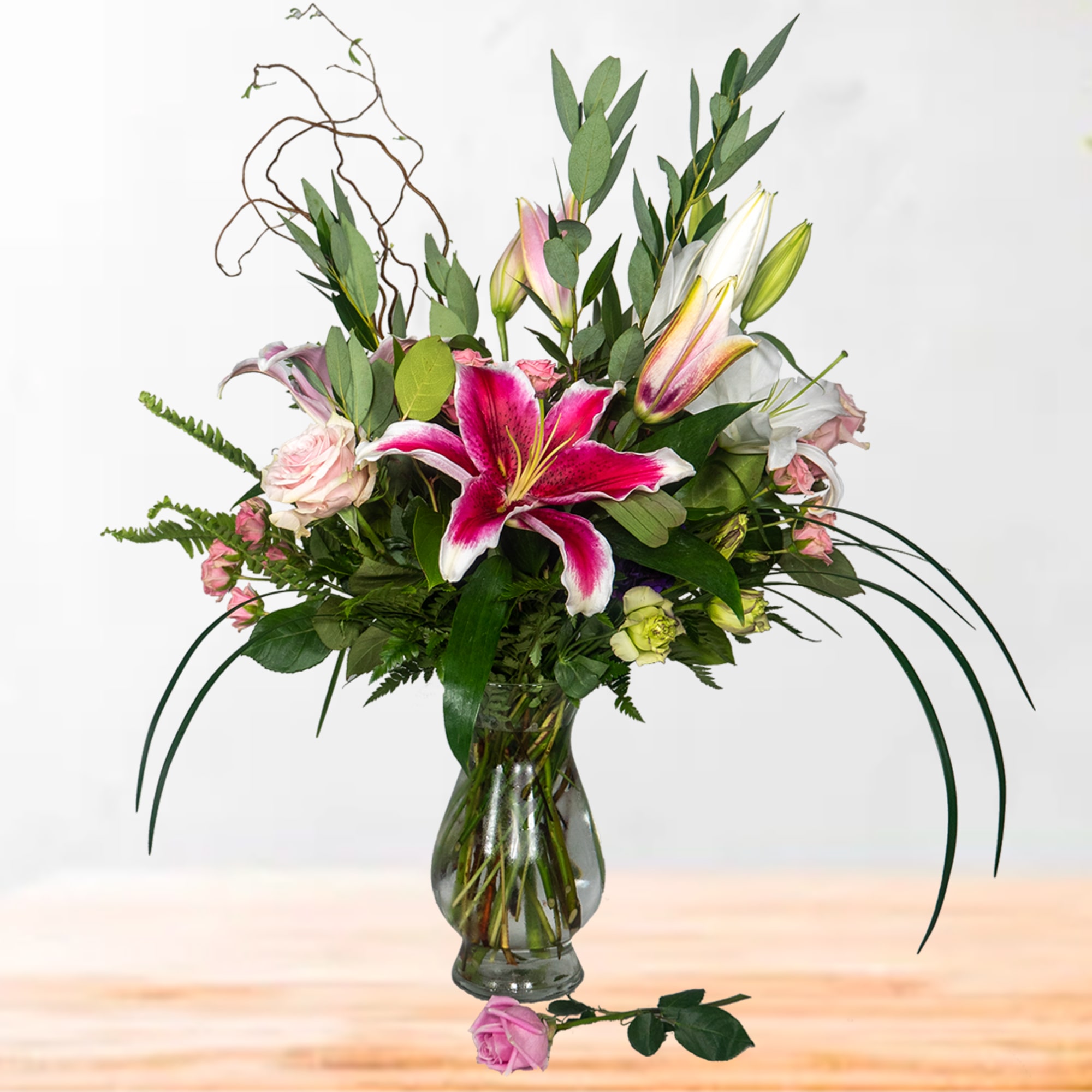 Tall bouquet of stargazer lilies and pink roses in a clear glass vase with a single rose on the table.