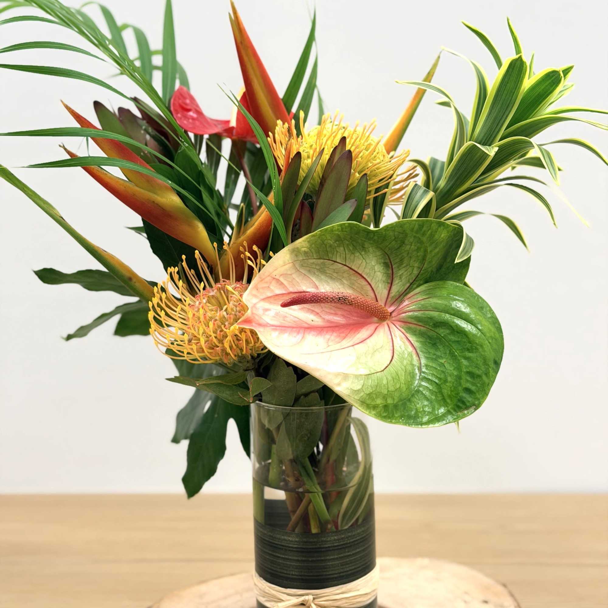 Tall tropical arrangement with anthurium, pincushion protea, and palm fronds in a clear glass vase.