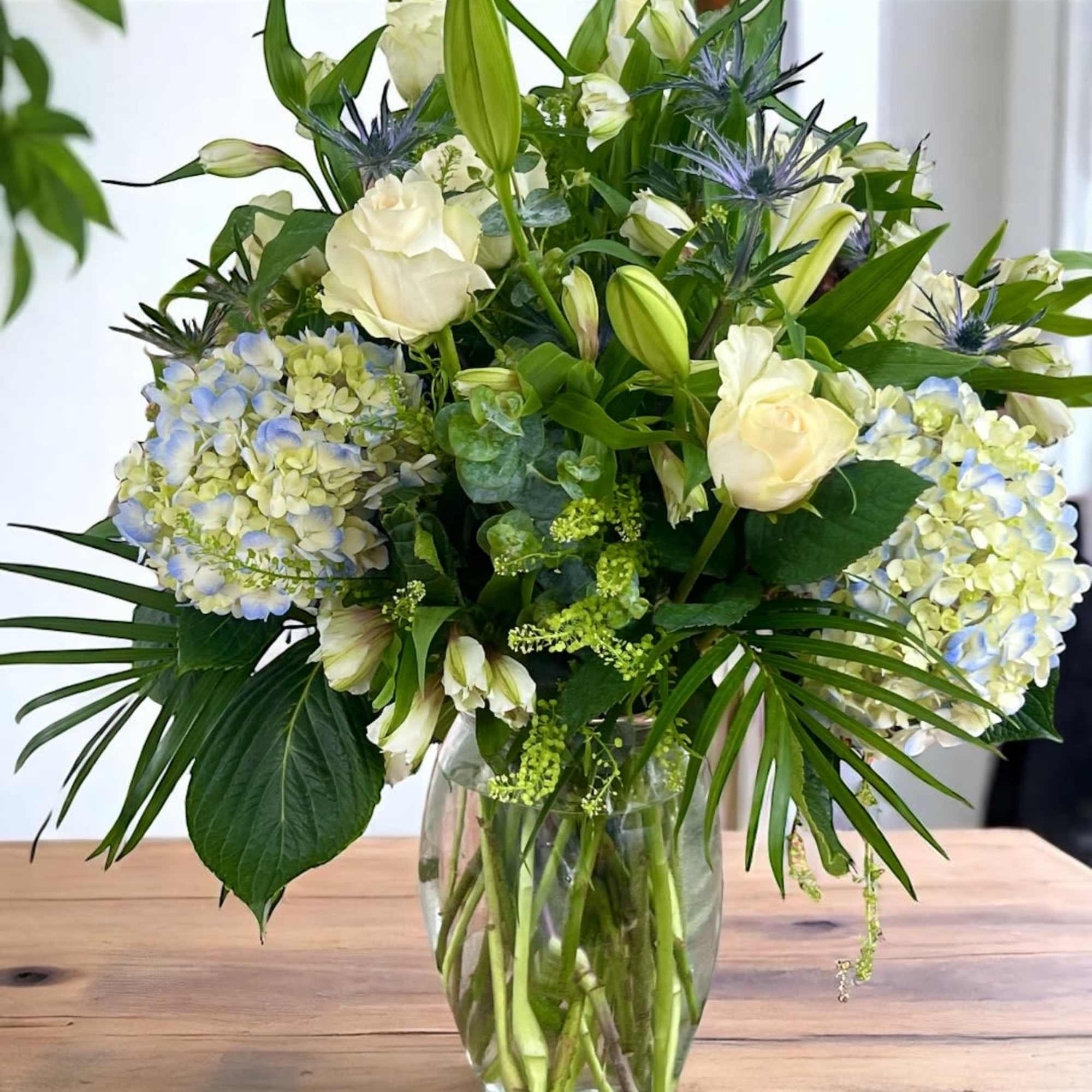 White roses and lilies arranged in a clear glass vase with blue hydrangeas