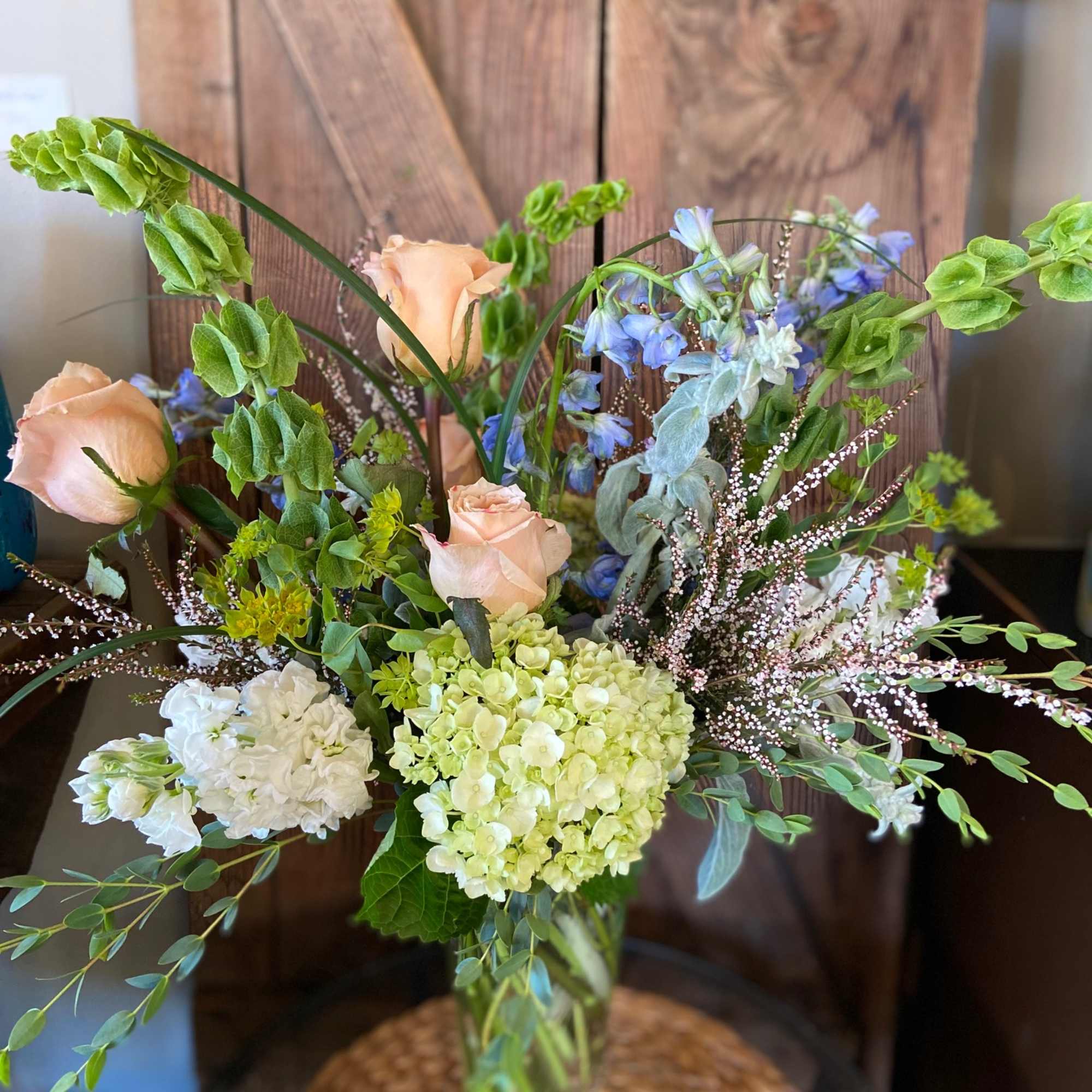Tall vase arrangement with peach roses, green hydrangeas, bells of Ireland, and mixed accent flowers