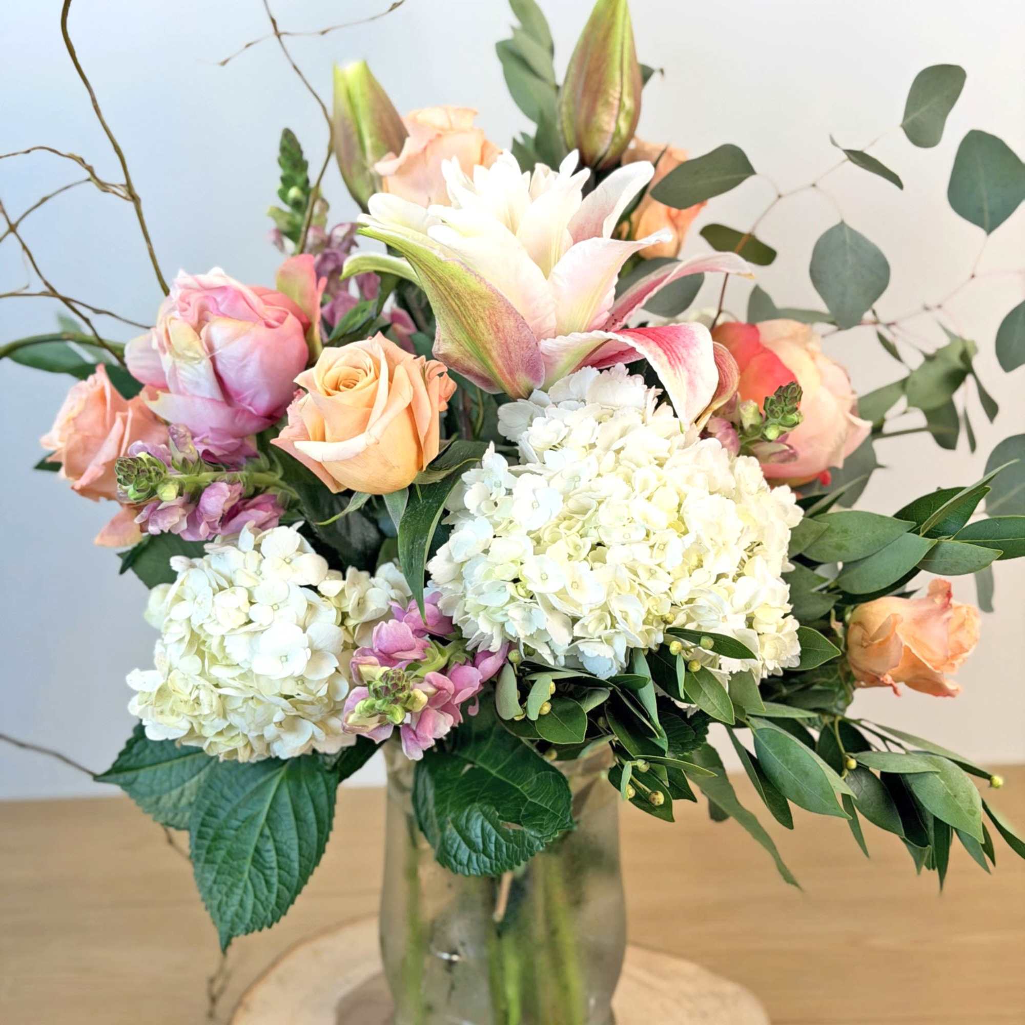 Mixed bouquet of peach roses, white hydrangeas, and pink lilies in a clear vase on a wood slice