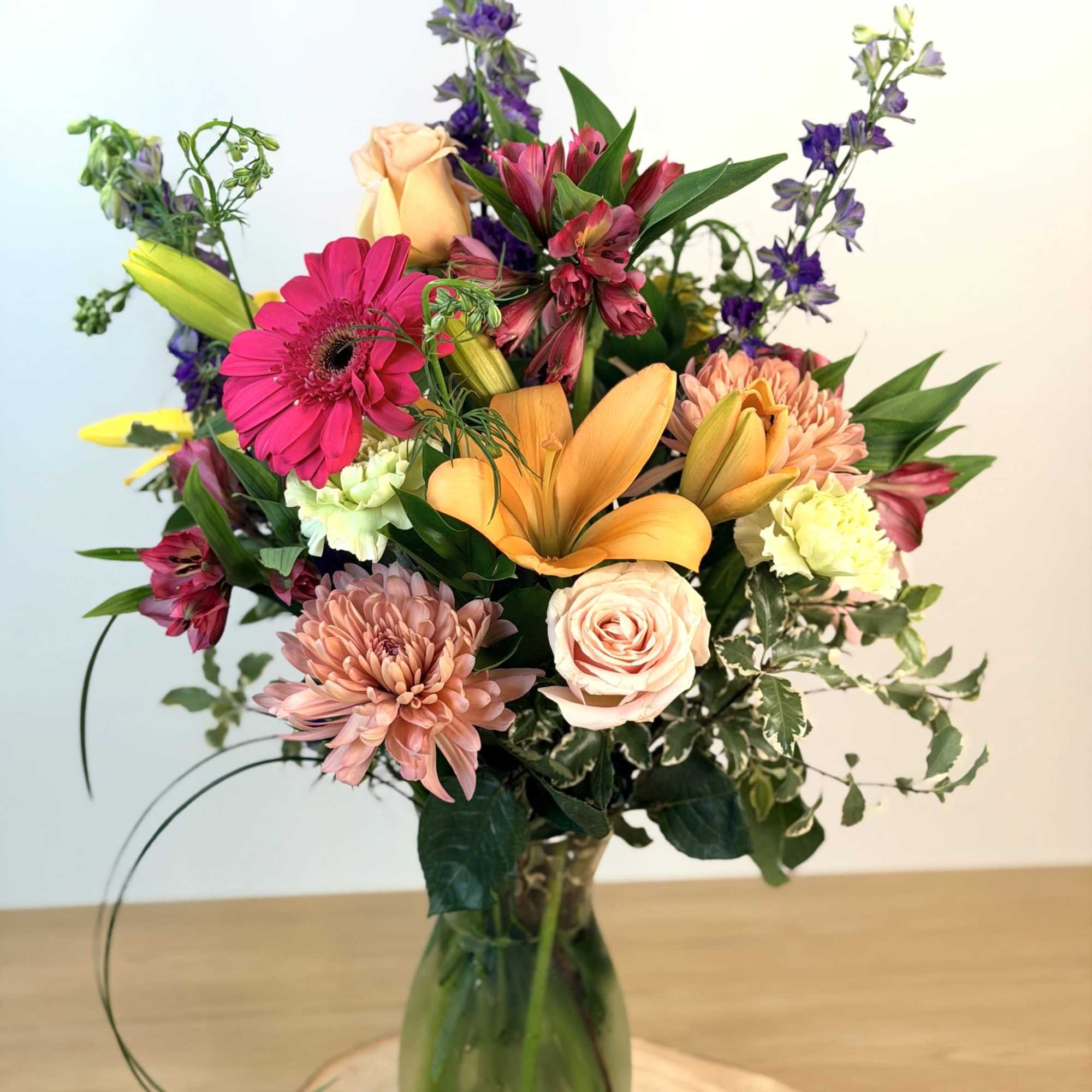 Tall mixed bouquet of pink, peach, yellow, and purple flowers in a clear glass vase on a wood base.