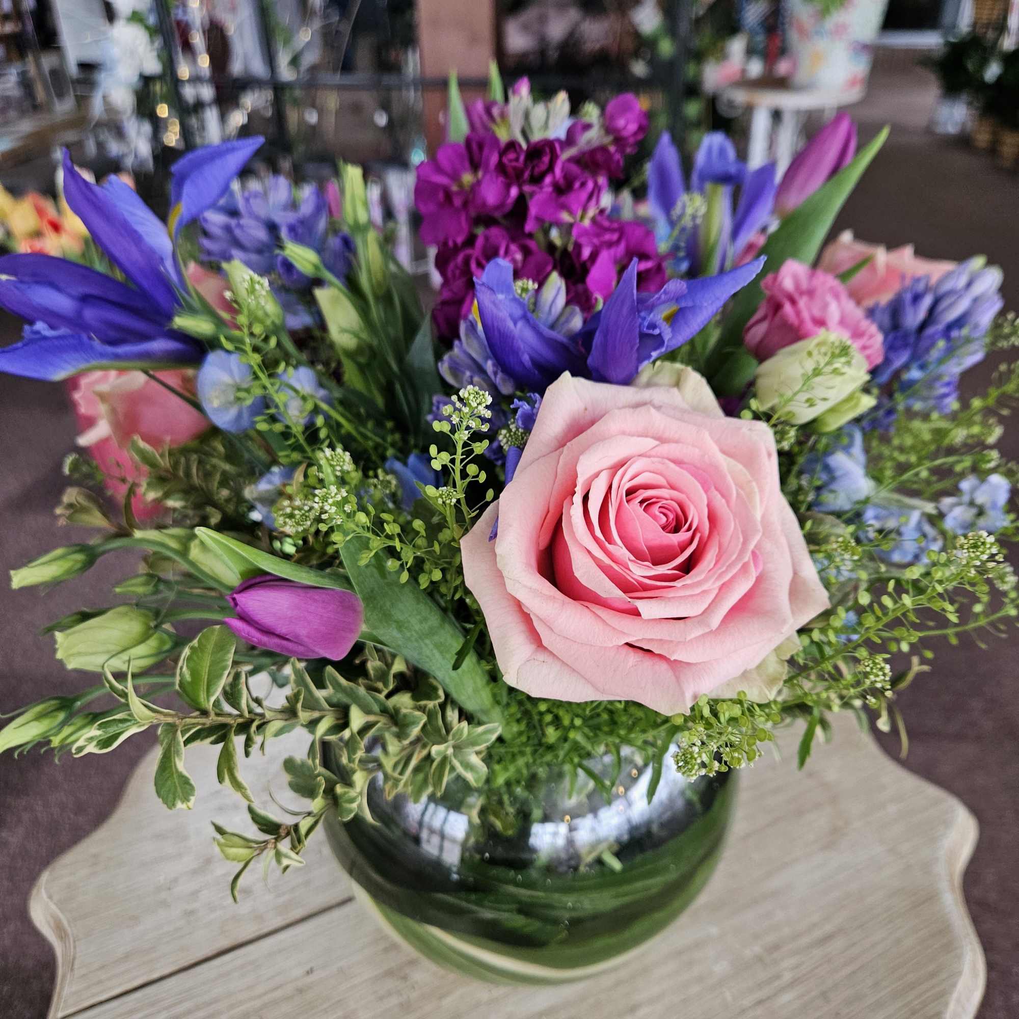 Low glass bowl arrangement with pink roses, purple stock, and blue irises.