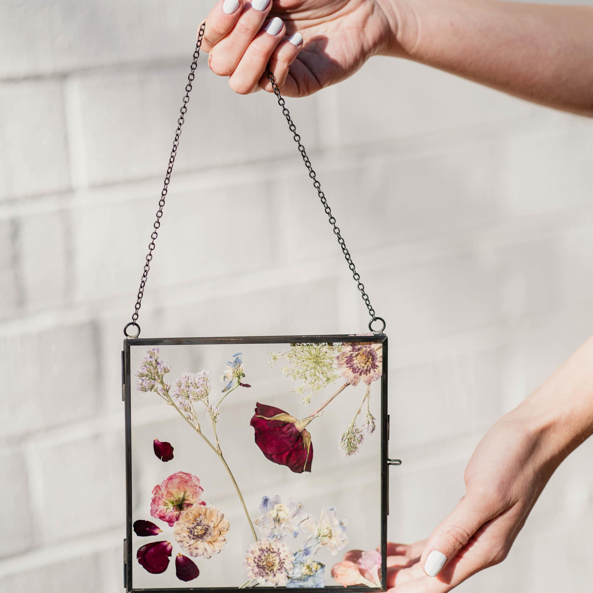 Hands holding a framed pressed-flower display with a hanging chain