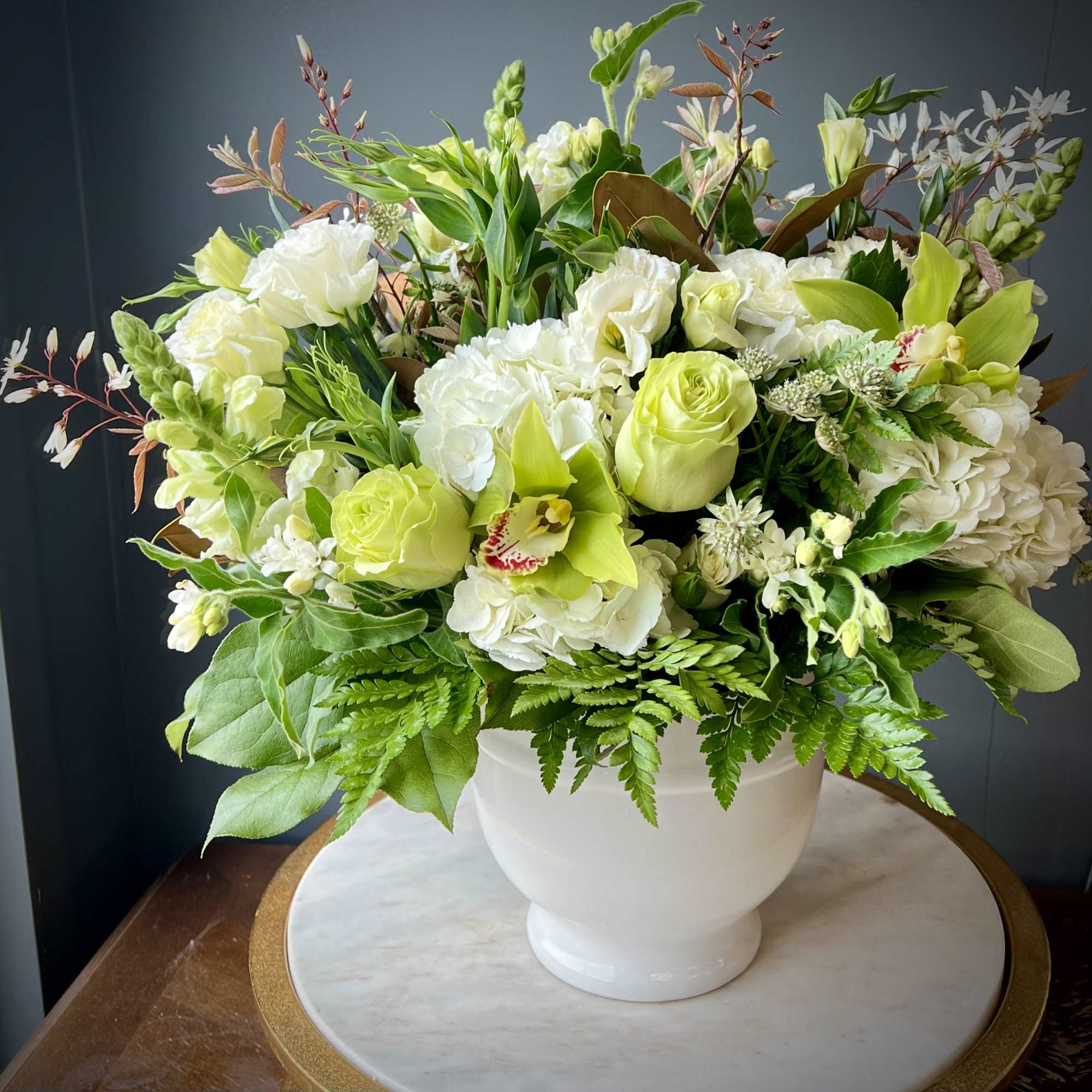 Low arrangement of white hydrangeas, green roses, and green orchids in a white bowl vase.