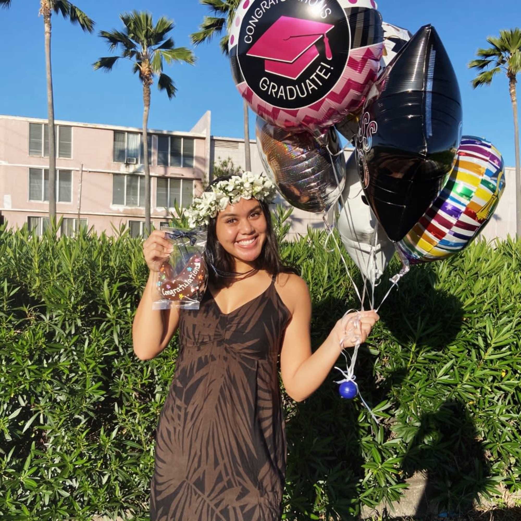 Young woman wearing a white flower crown holding graduation balloons and a cookie gift