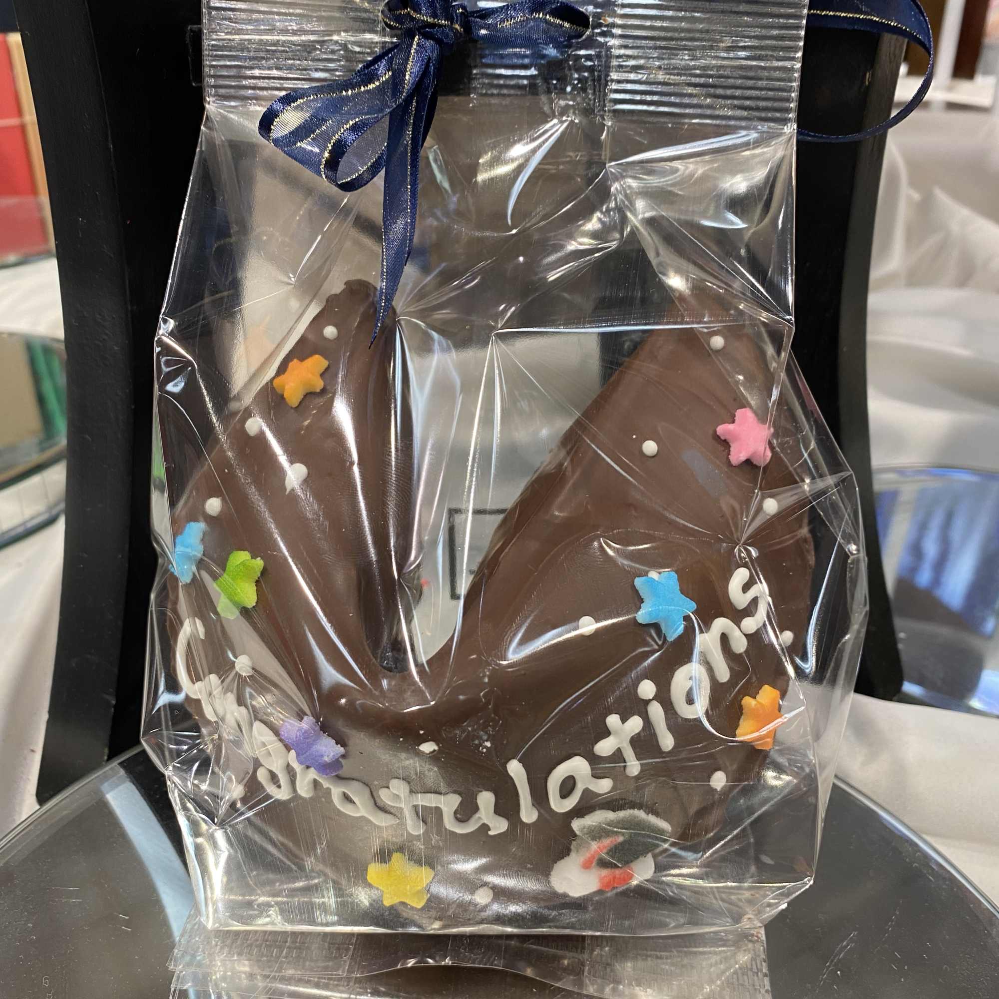 Large chocolate fortune cookie with sprinkles and congratulations text wrapped in clear cellophane with a blue ribbon.