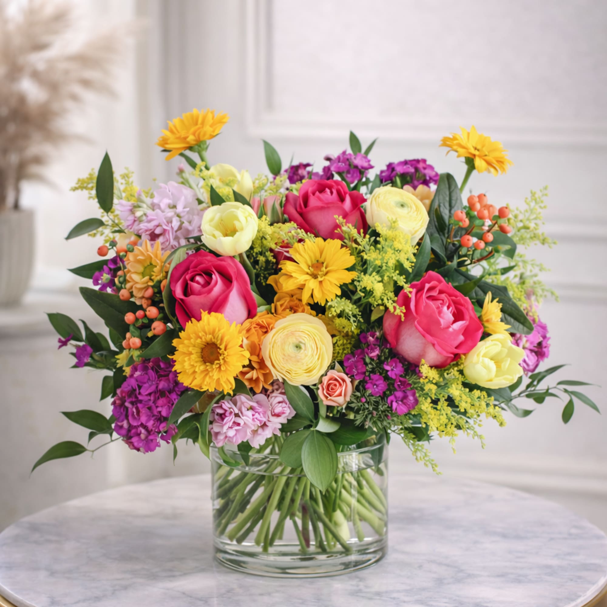 Bright mixed bouquet of pink roses, yellow ranunculus and daisies in a clear glass vase.