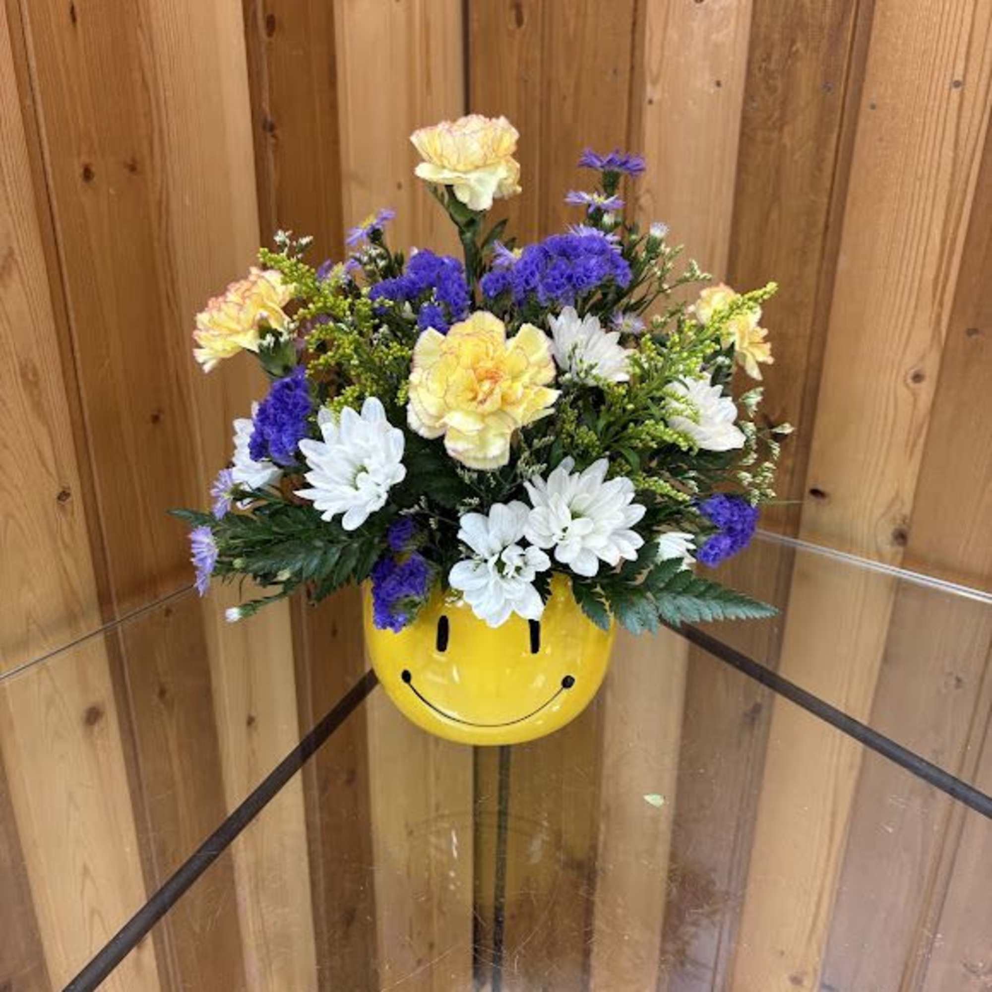 Cheerful mixed bouquet of yellow carnations, white mums, and purple blooms in a yellow smiley face container.