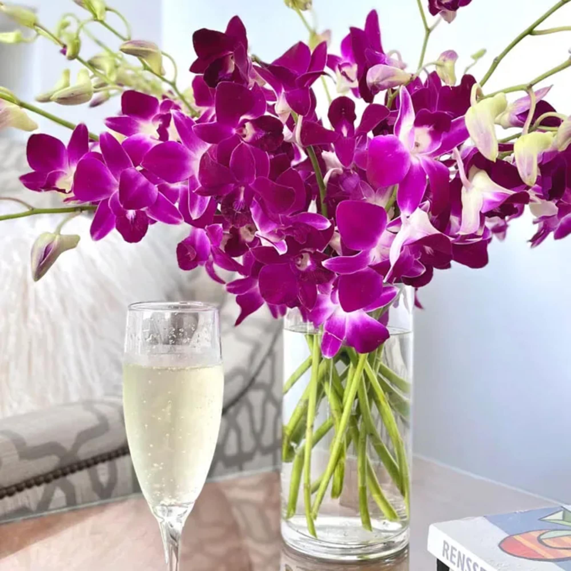 Purple orchids in a clear glass vase beside a champagne flute