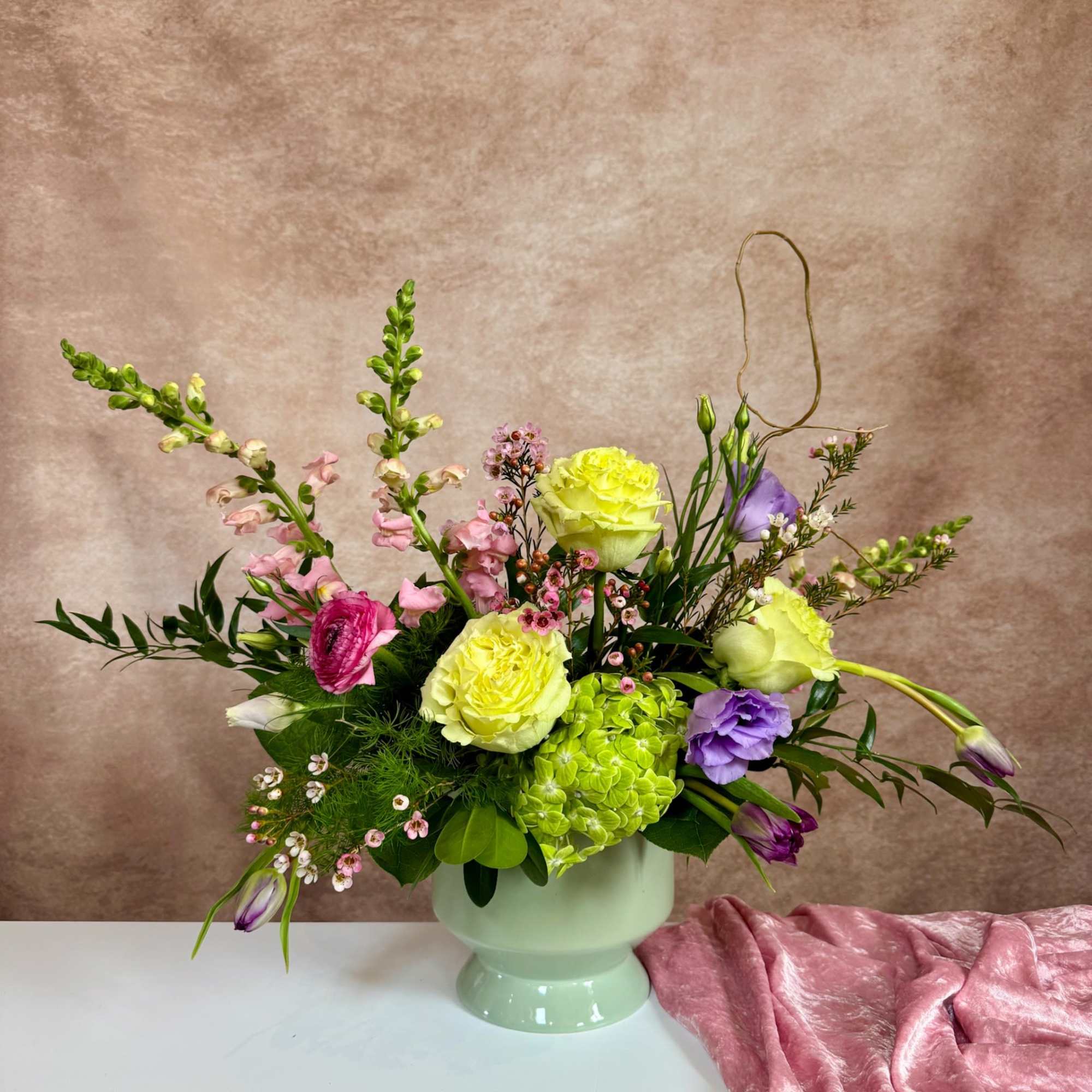Asymmetrical pastel arrangement with yellow roses, green hydrangea and mixed blooms in a pale green pedestal vase