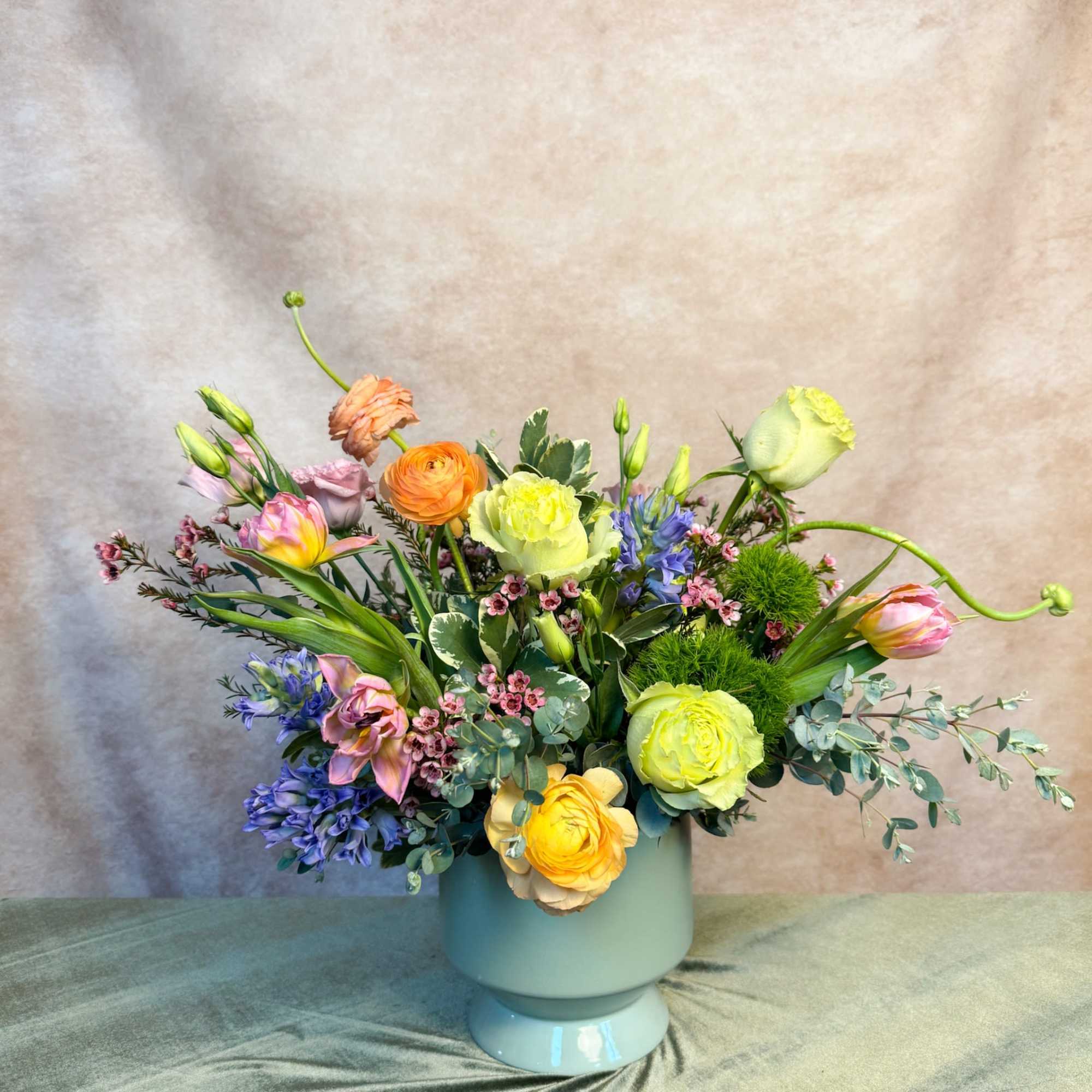 Low pastel arrangement of roses, ranunculus, tulips and mixed blooms in a light blue pedestal vase
