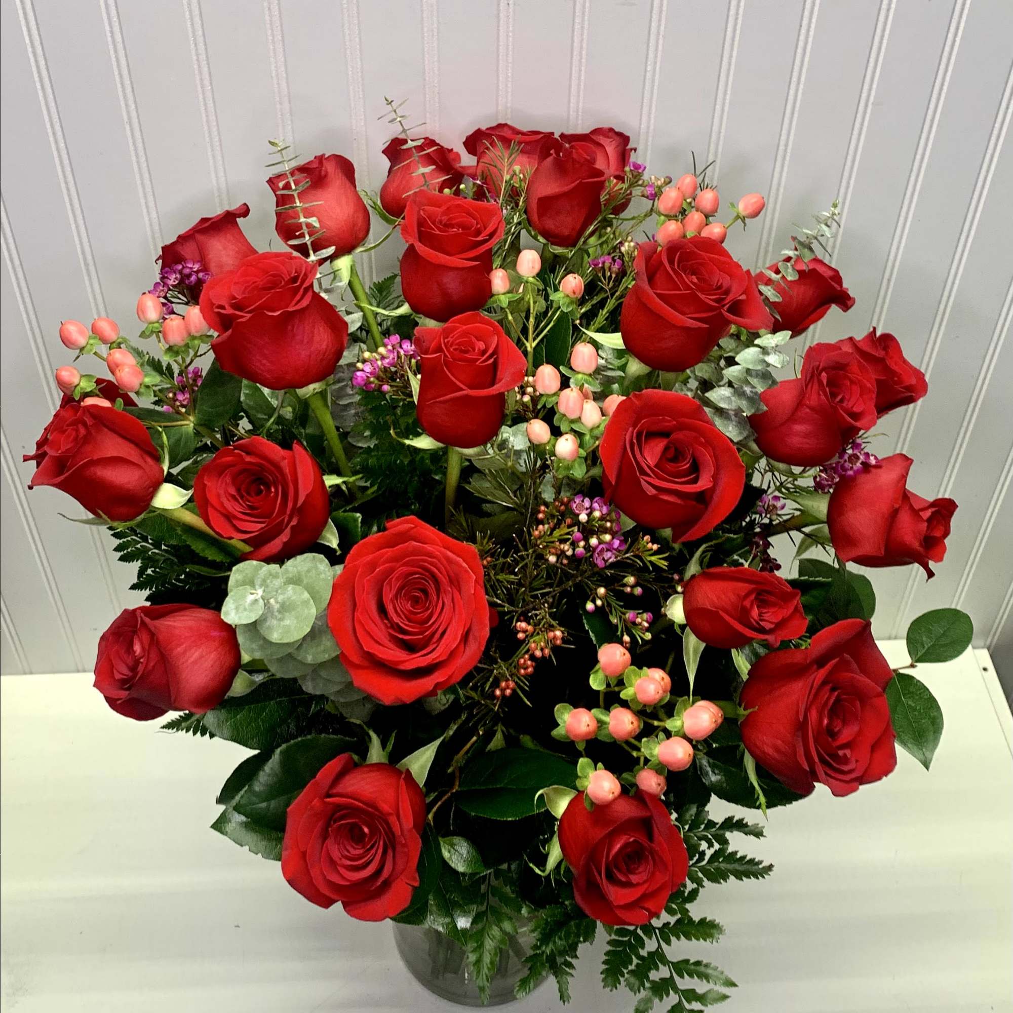 Tall arrangement of red roses with pink berries and purple filler flowers in a clear glass vase