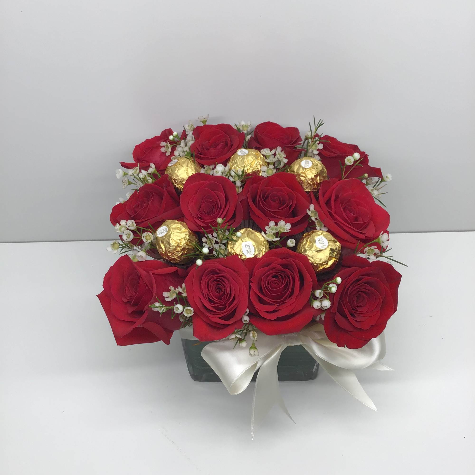 Low arrangement of red roses with gold-wrapped chocolates in a clear cube vase with white bow