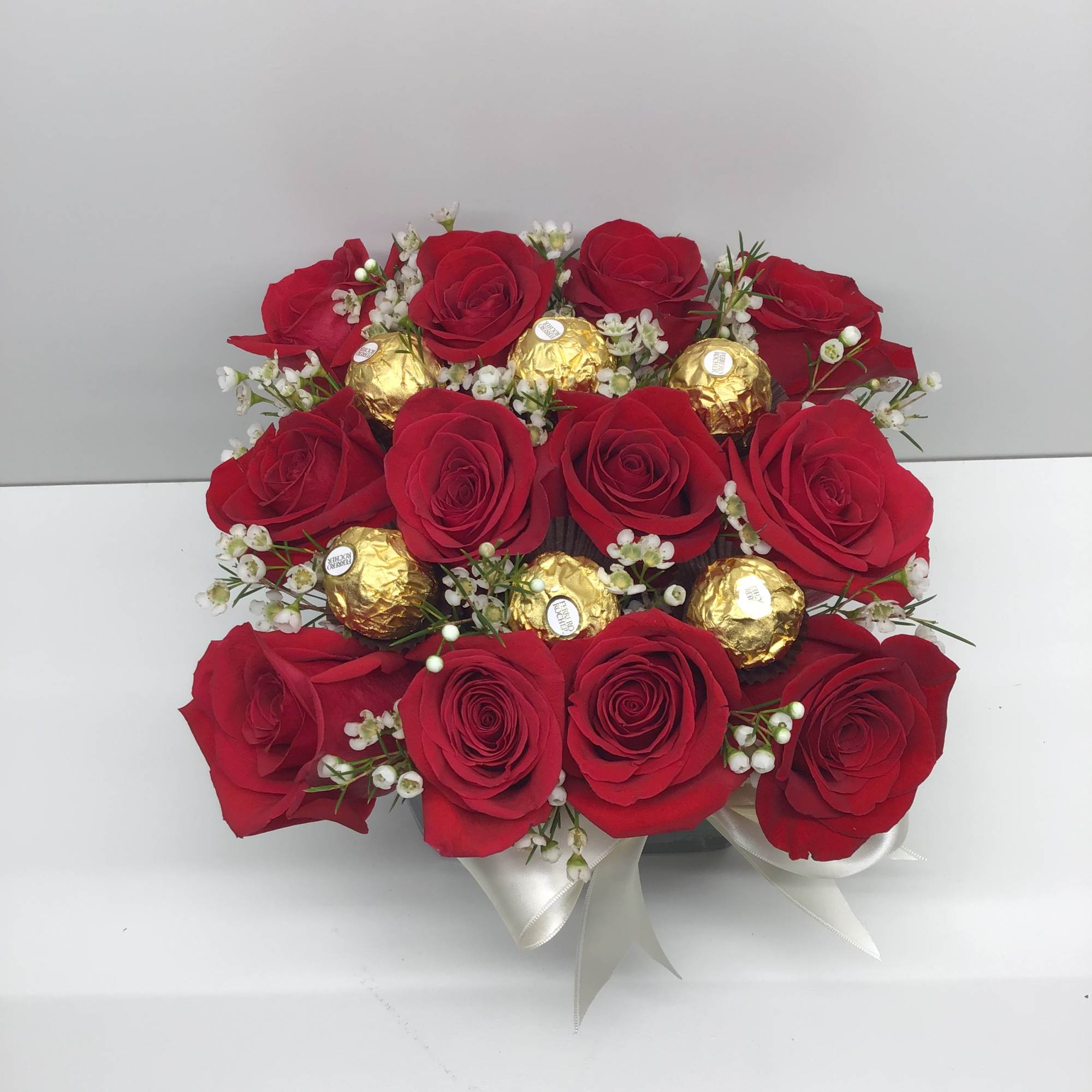 Square arrangement of red roses with gold-wrapped chocolates and small white blooms in a box with ribbon