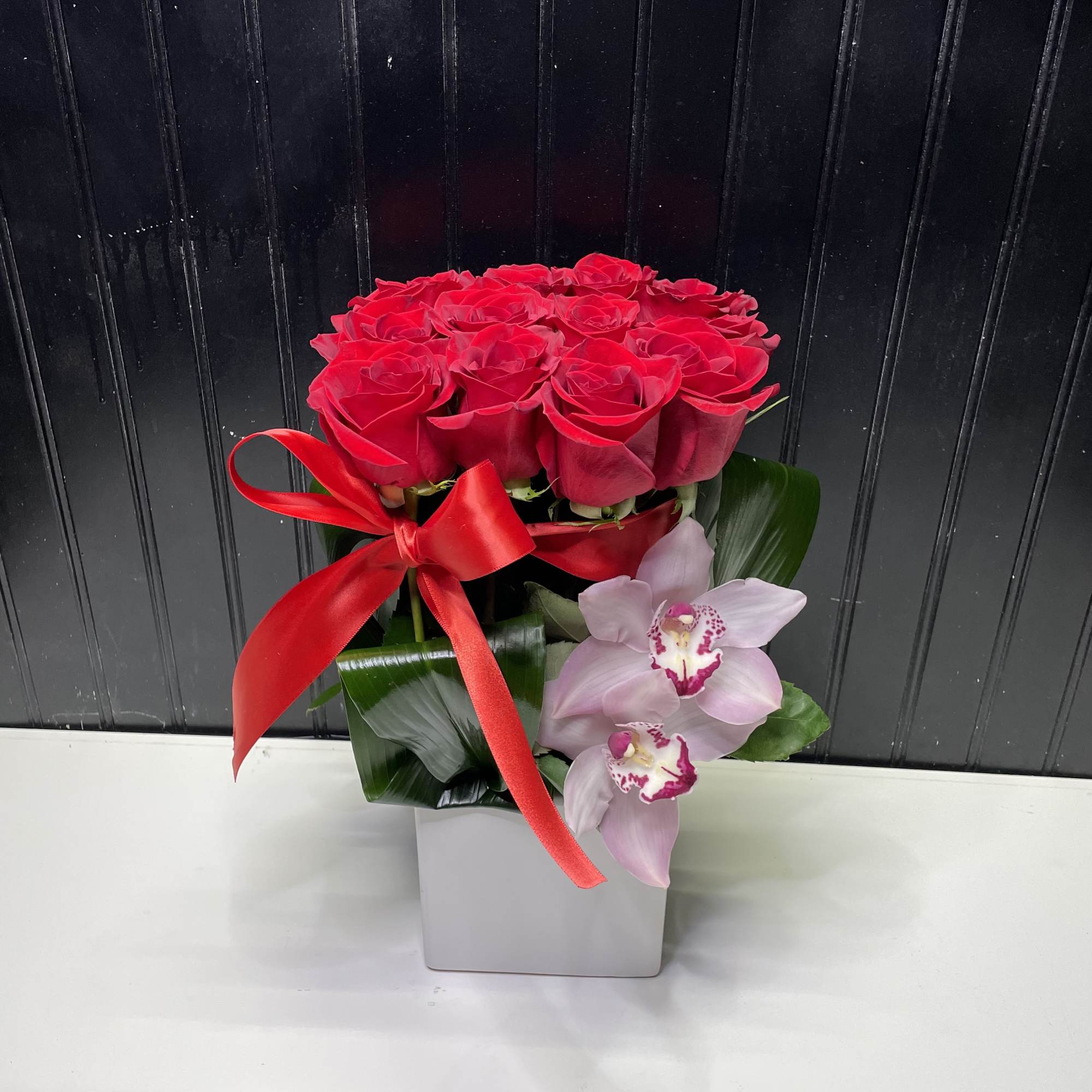 Compact arrangement of red roses with pink orchids in a square white vase with a red ribbon bow