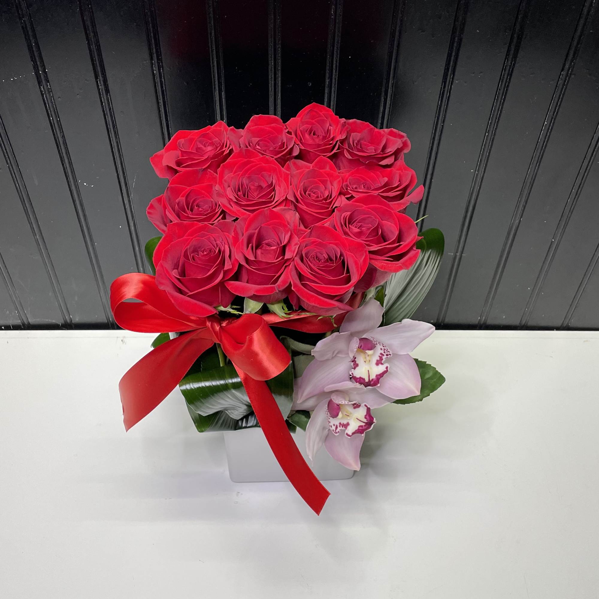 Compact arrangement of red roses with two pink orchids in a white cube vase tied with a red ribbon