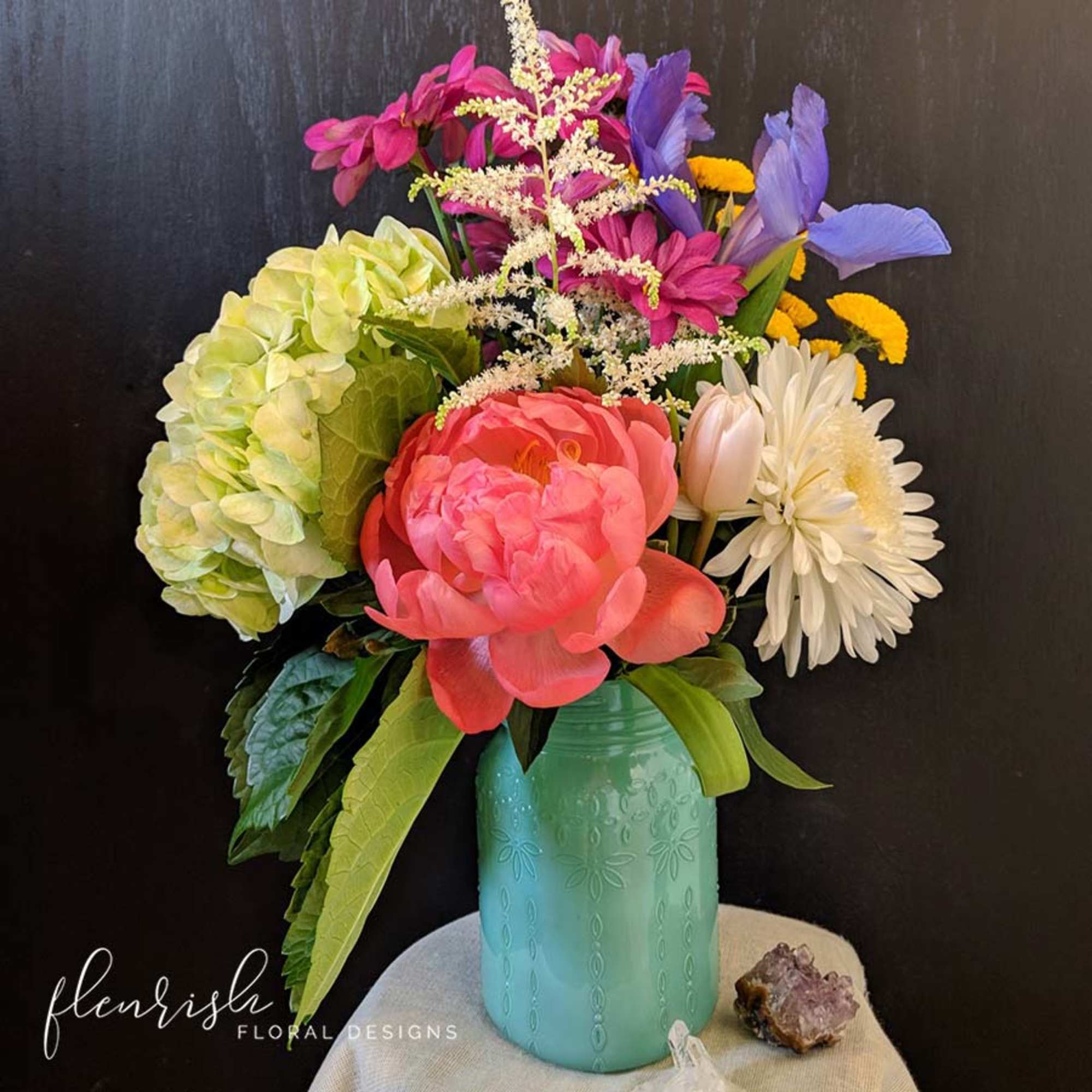Colorful mixed bouquet with peony, hydrangea, iris and daisies in a teal jar-style vase