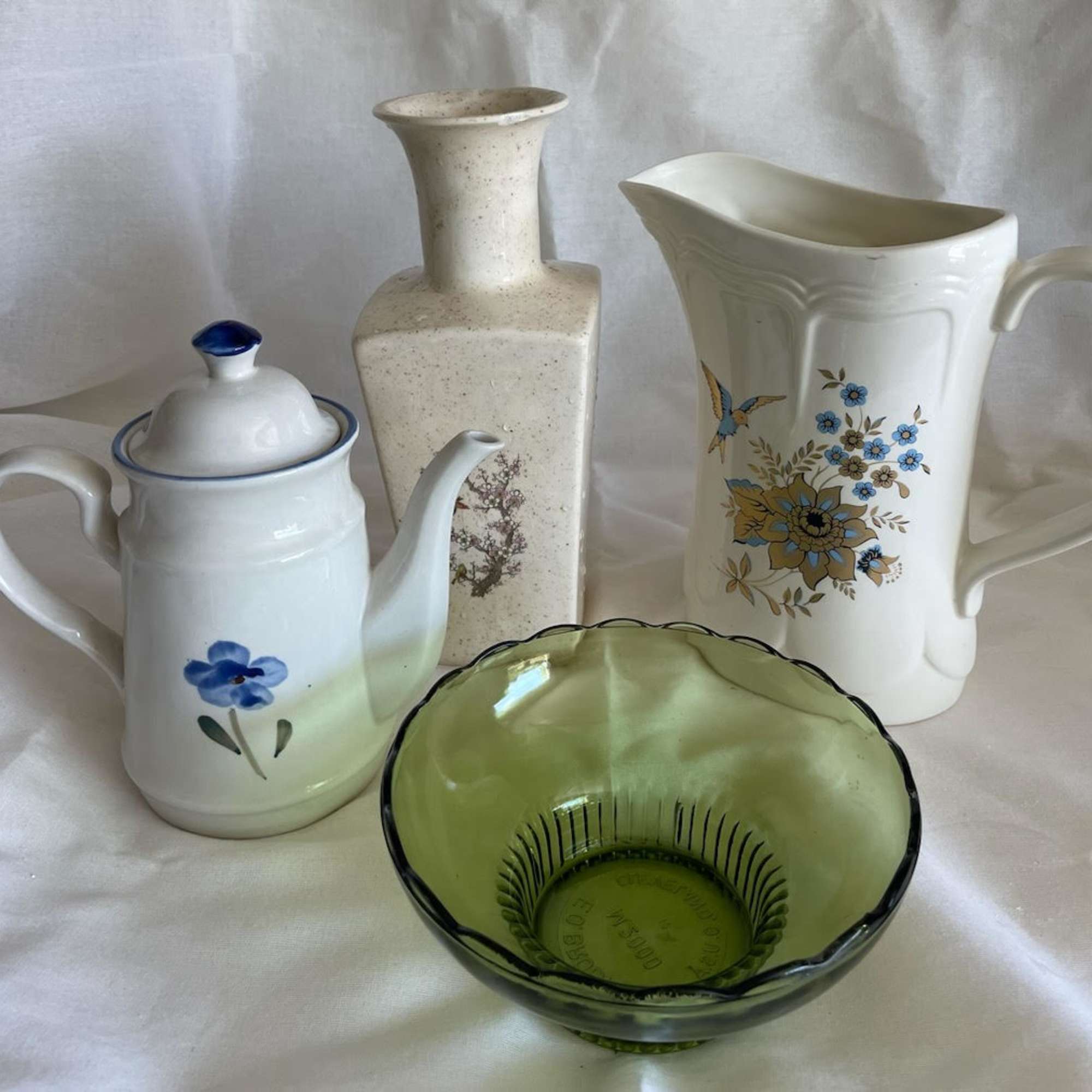 Set of vintage-style ceramic pitchers, teapot, vase, and green glass bowl arranged on fabric.