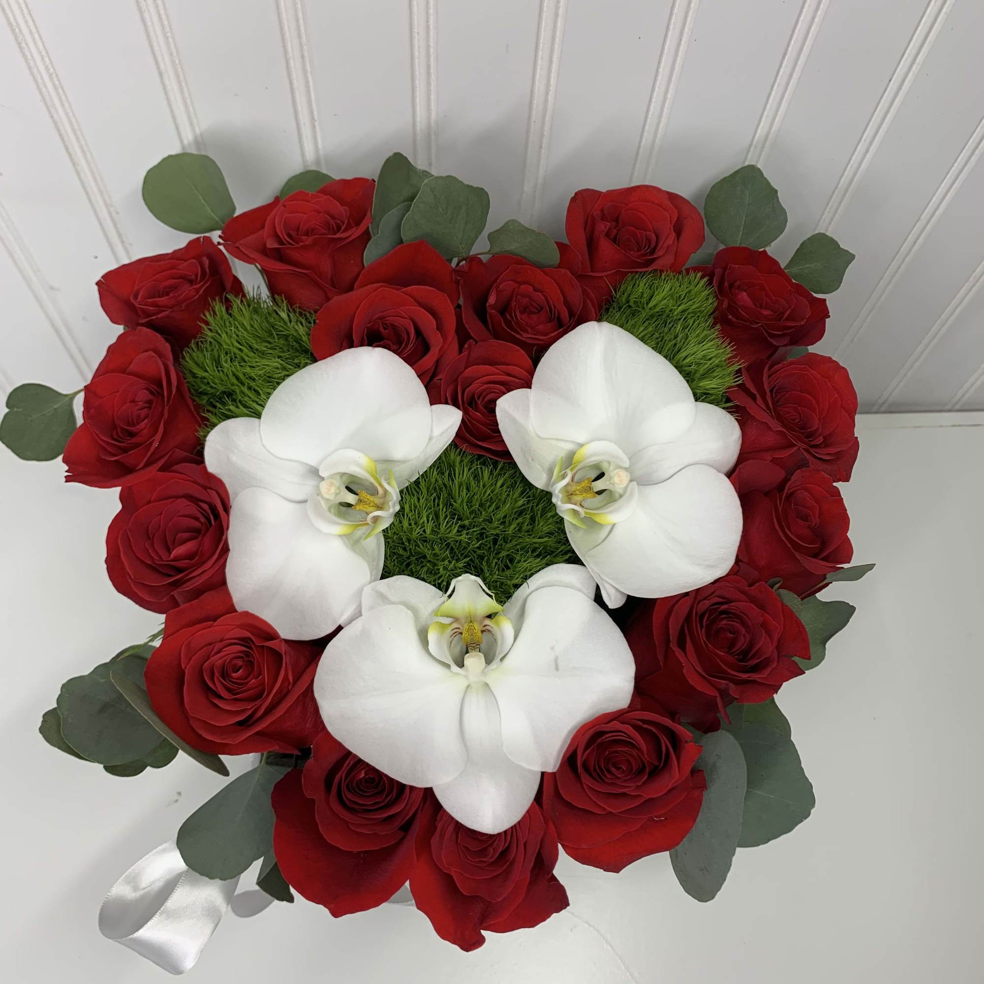 Heart-shaped arrangement of red roses with white orchids and green moss accents.