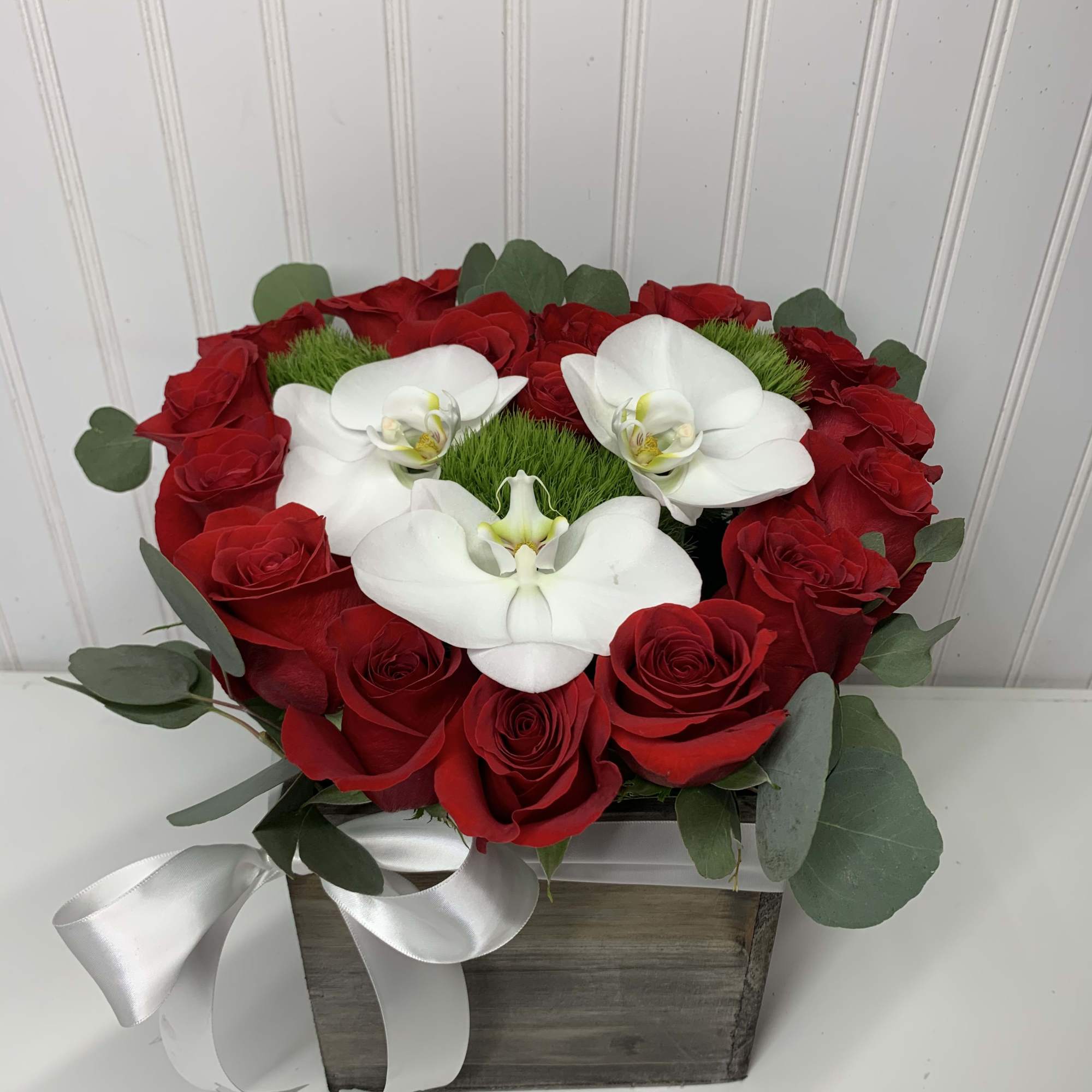 Heart-shaped arrangement of red roses and white orchids in a wood box with white ribbon