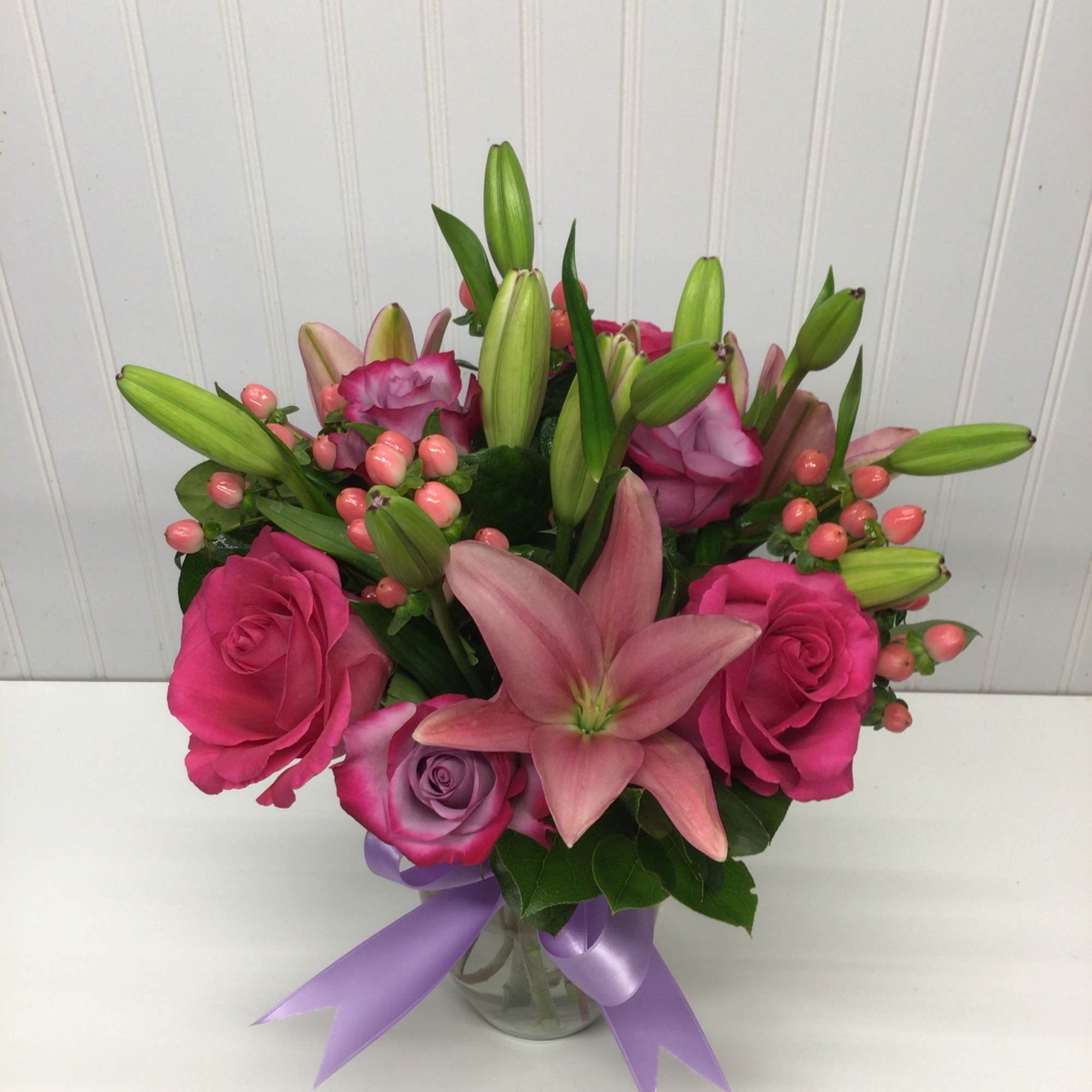 Pink roses and lilies with berry accents in a clear glass vase tied with a lavender ribbon