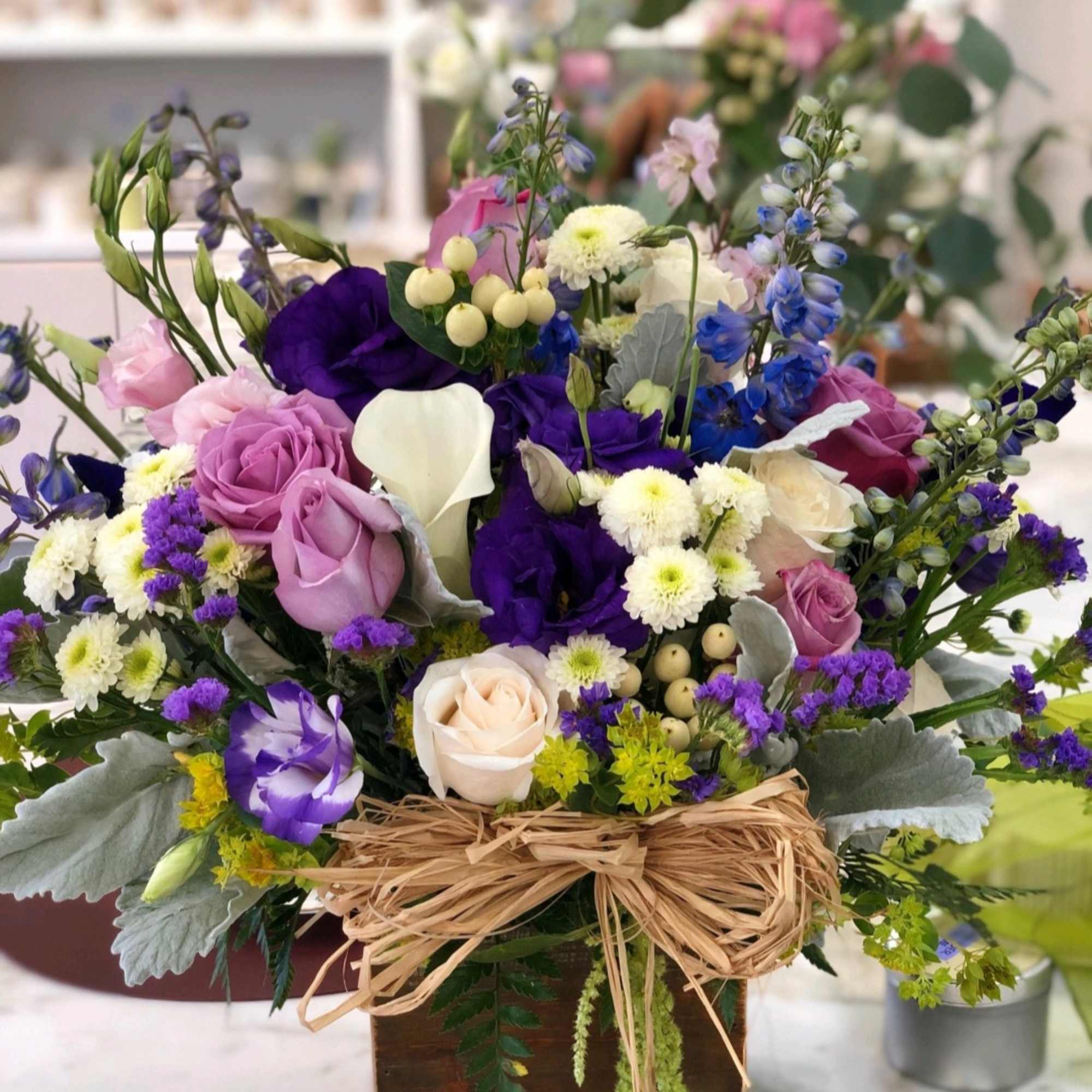 Mixed arrangement of purple, lavender, and white flowers in a wooden box with a raffia bow