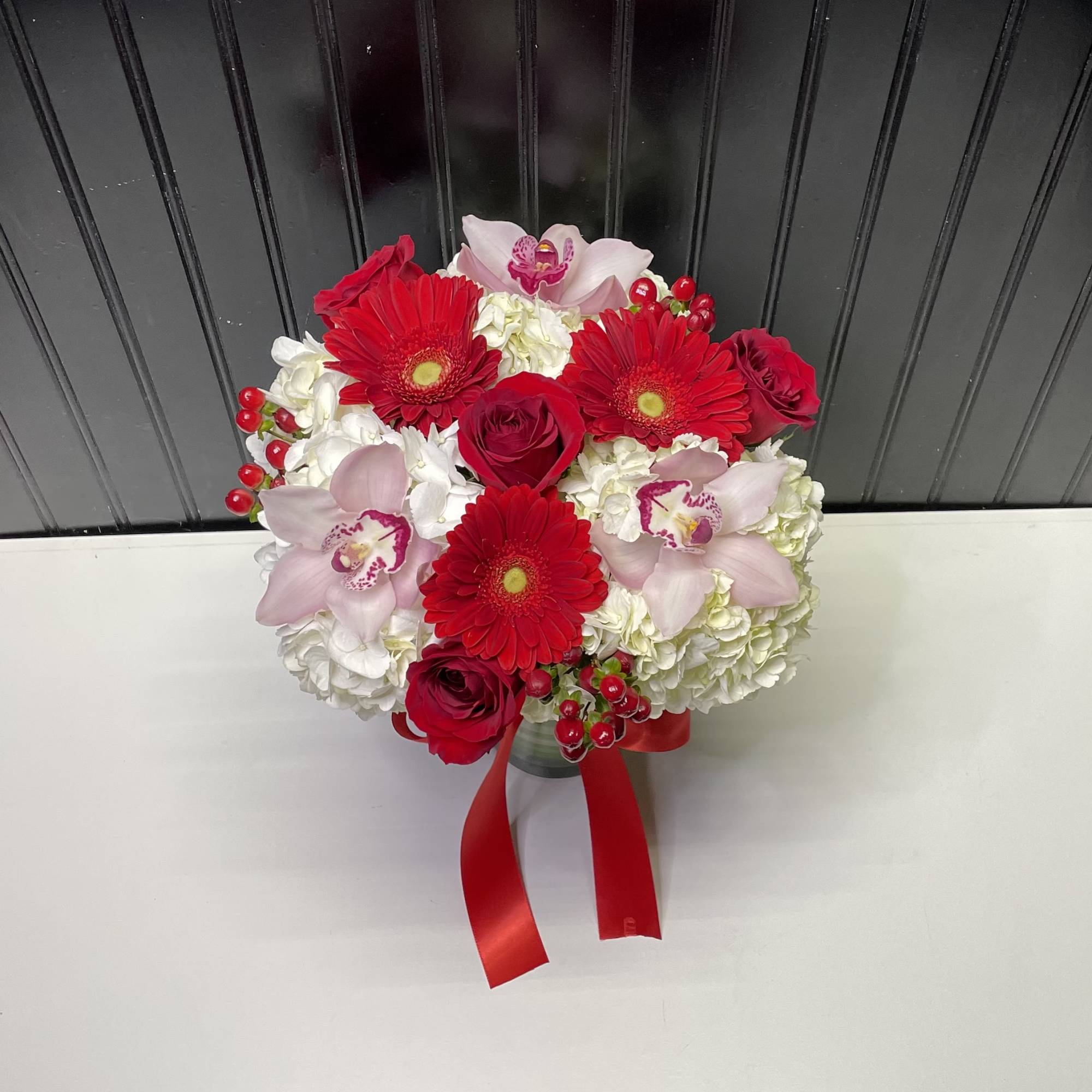 Compact red and white arrangement with gerbera daisies, roses, orchids, and hydrangeas in a vase with red ribbon