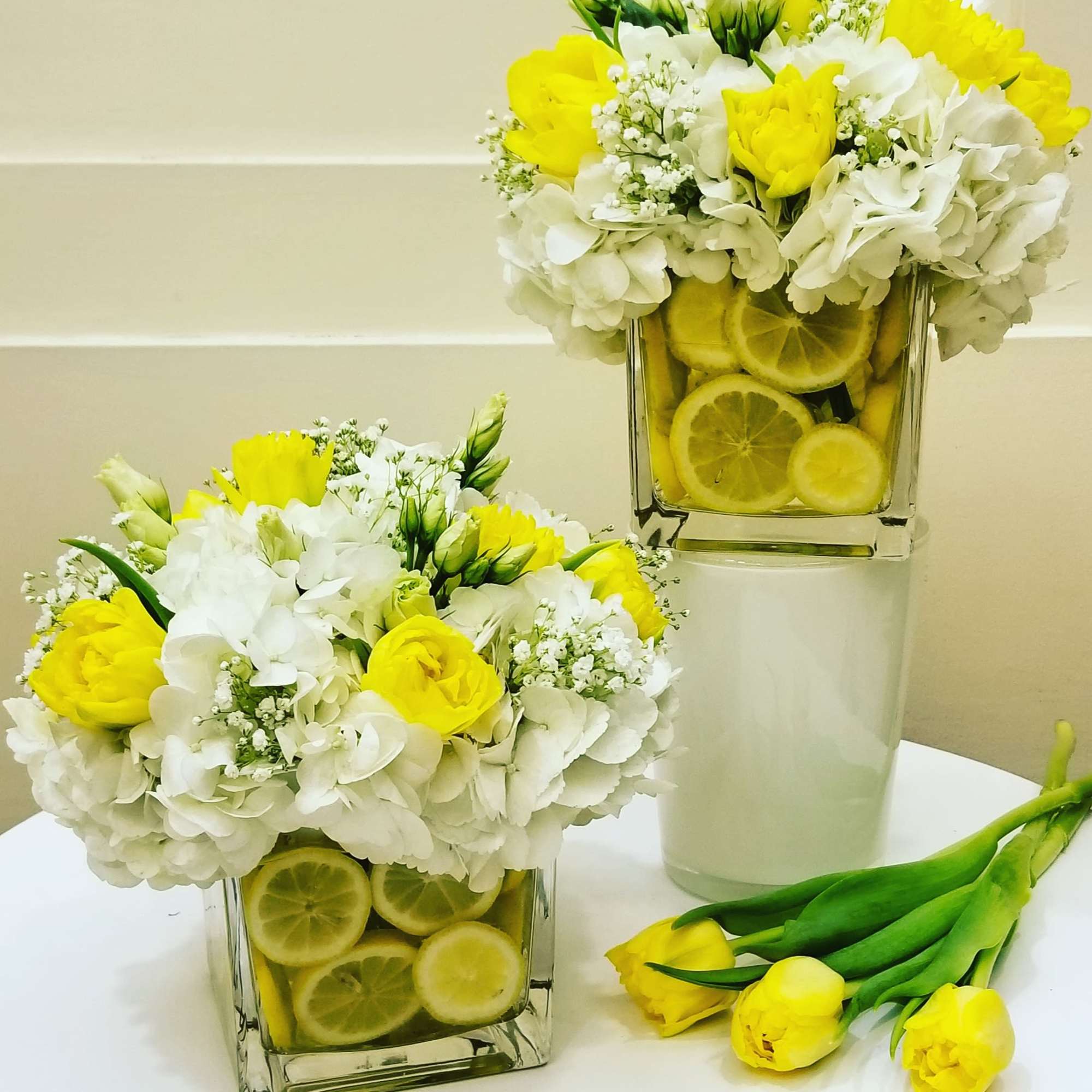Two yellow and white floral arrangements in square glass vases with lemon slices.