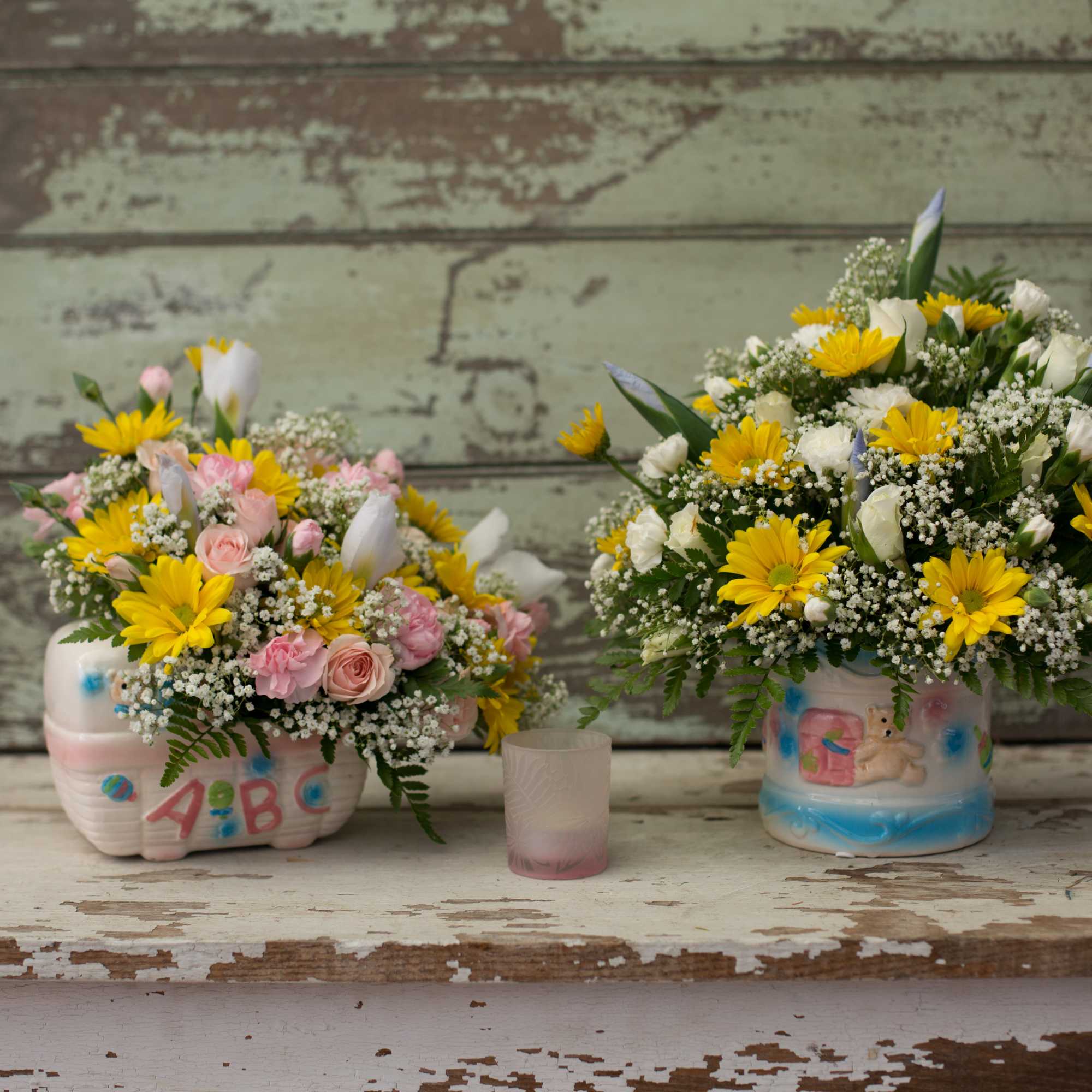 Two small floral arrangements in decorated containers with a candle between them