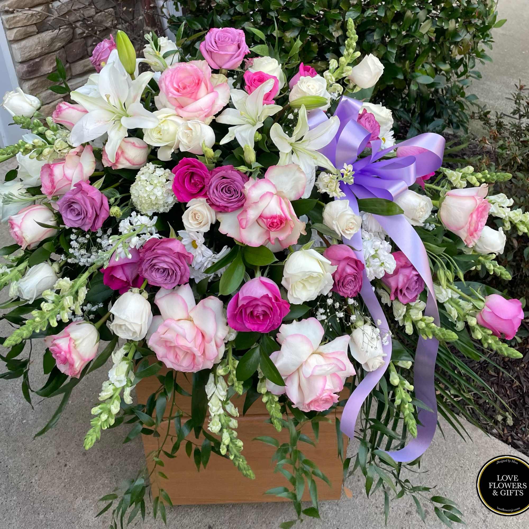 Large arrangement of pink, white, and lavender roses with white lilies and a lavender bow.