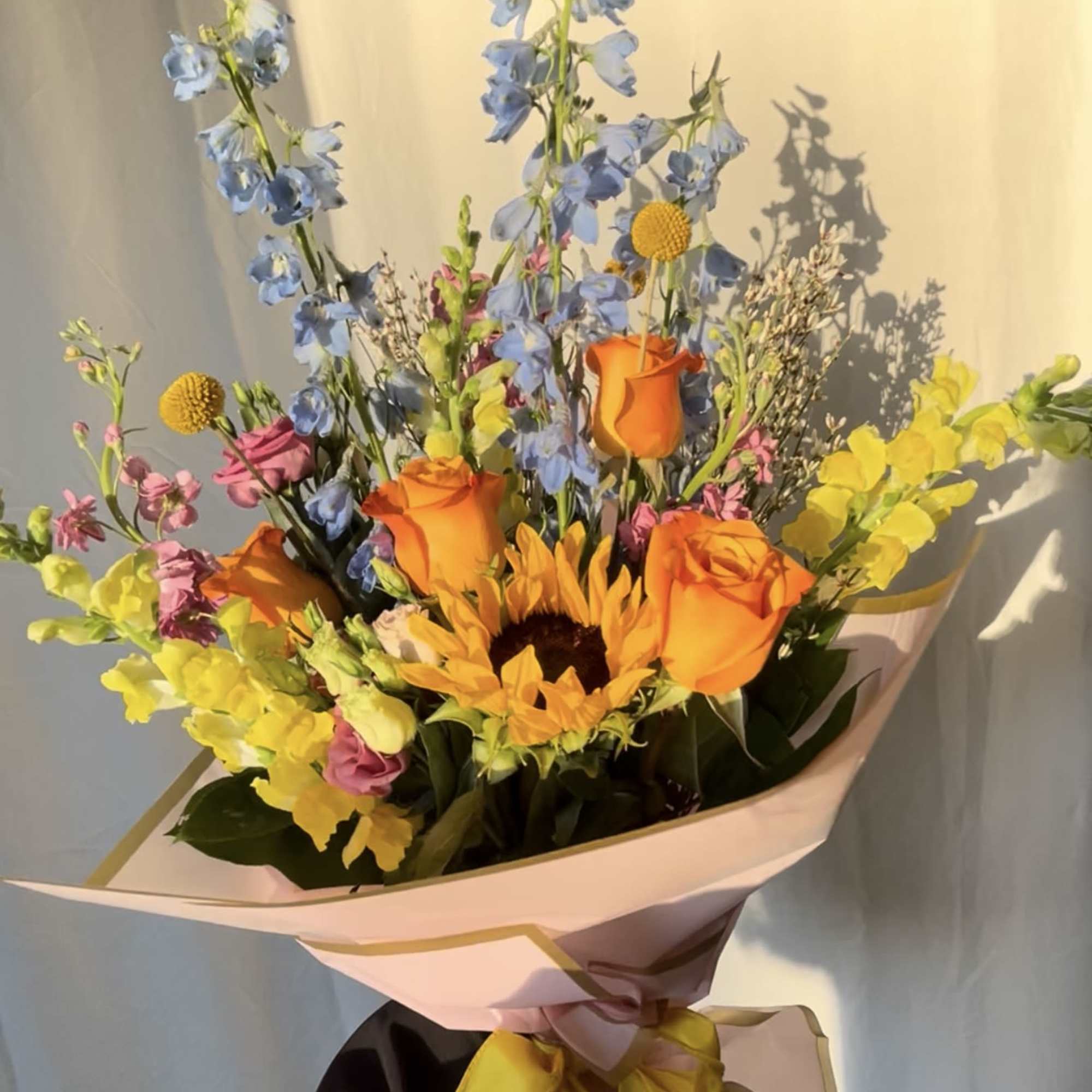 Bouquet of orange roses, blue delphinium, and a sunflower wrapped in pink paper