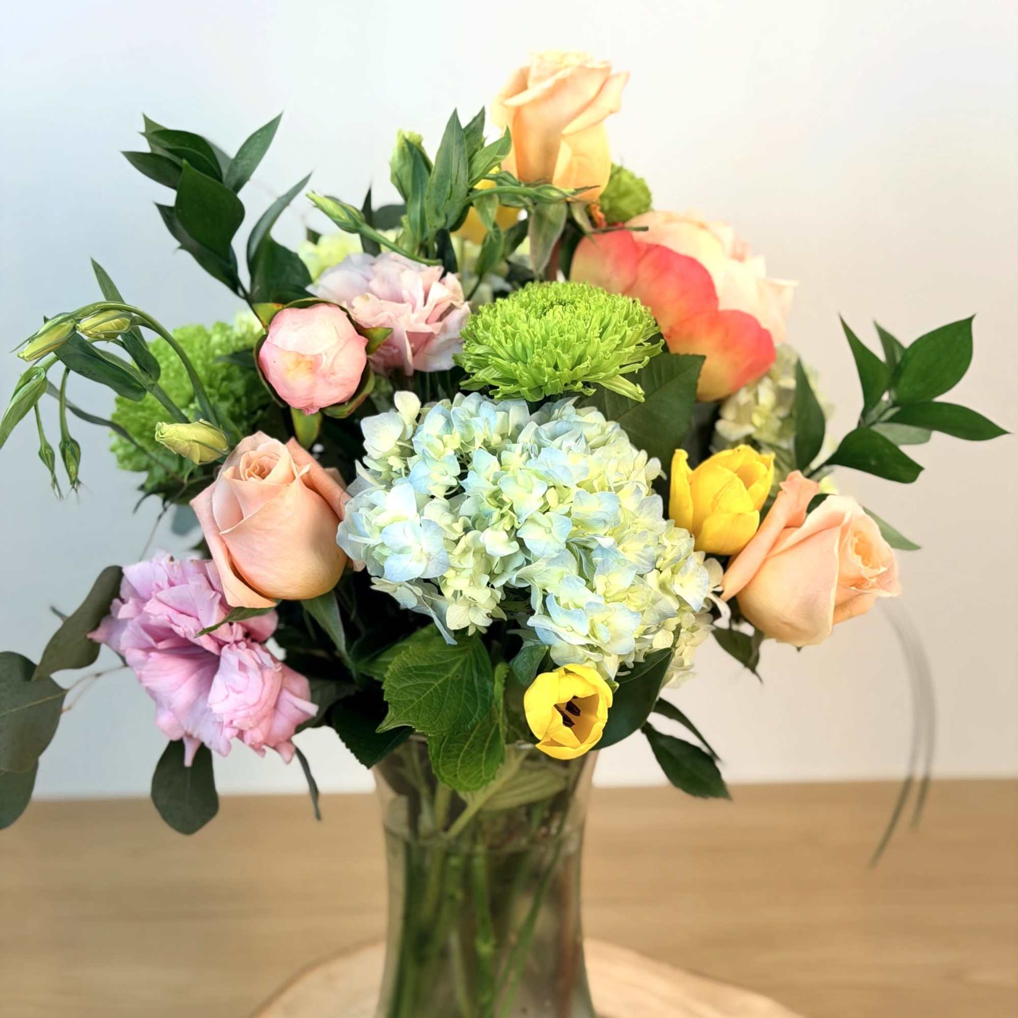 Bouquet of peach roses, yellow tulips, green mums, and blue hydrangeas in a clear glass vase