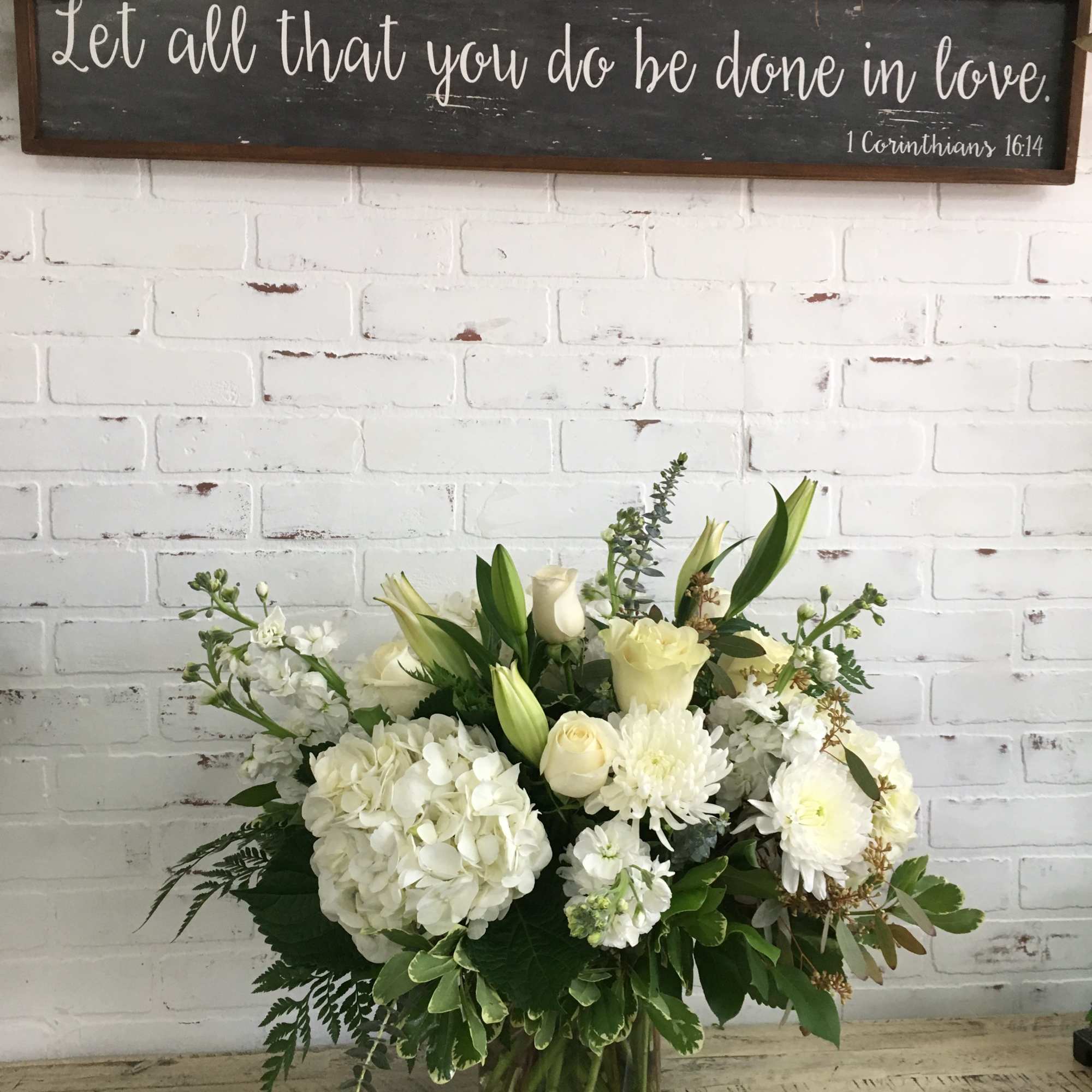 White hydrangeas, roses, lilies, and mums arranged in a clear glass vase against a white brick wall.
