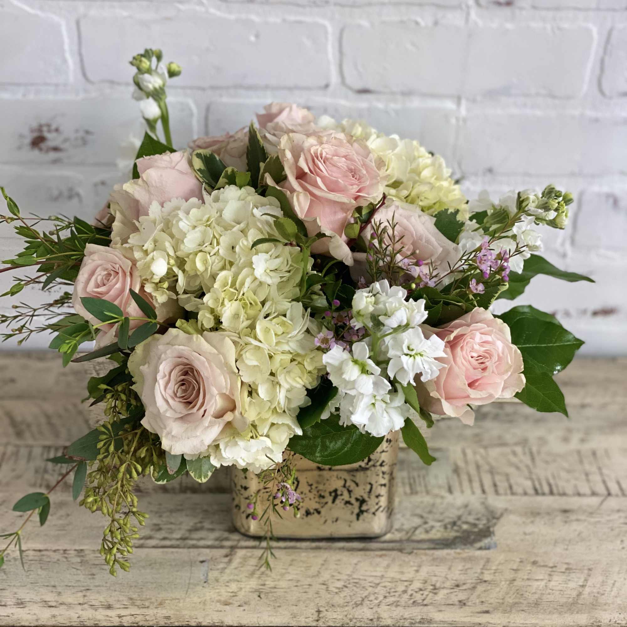 Low arrangement of blush roses and white hydrangeas in a gold cube vase on a rustic wood table