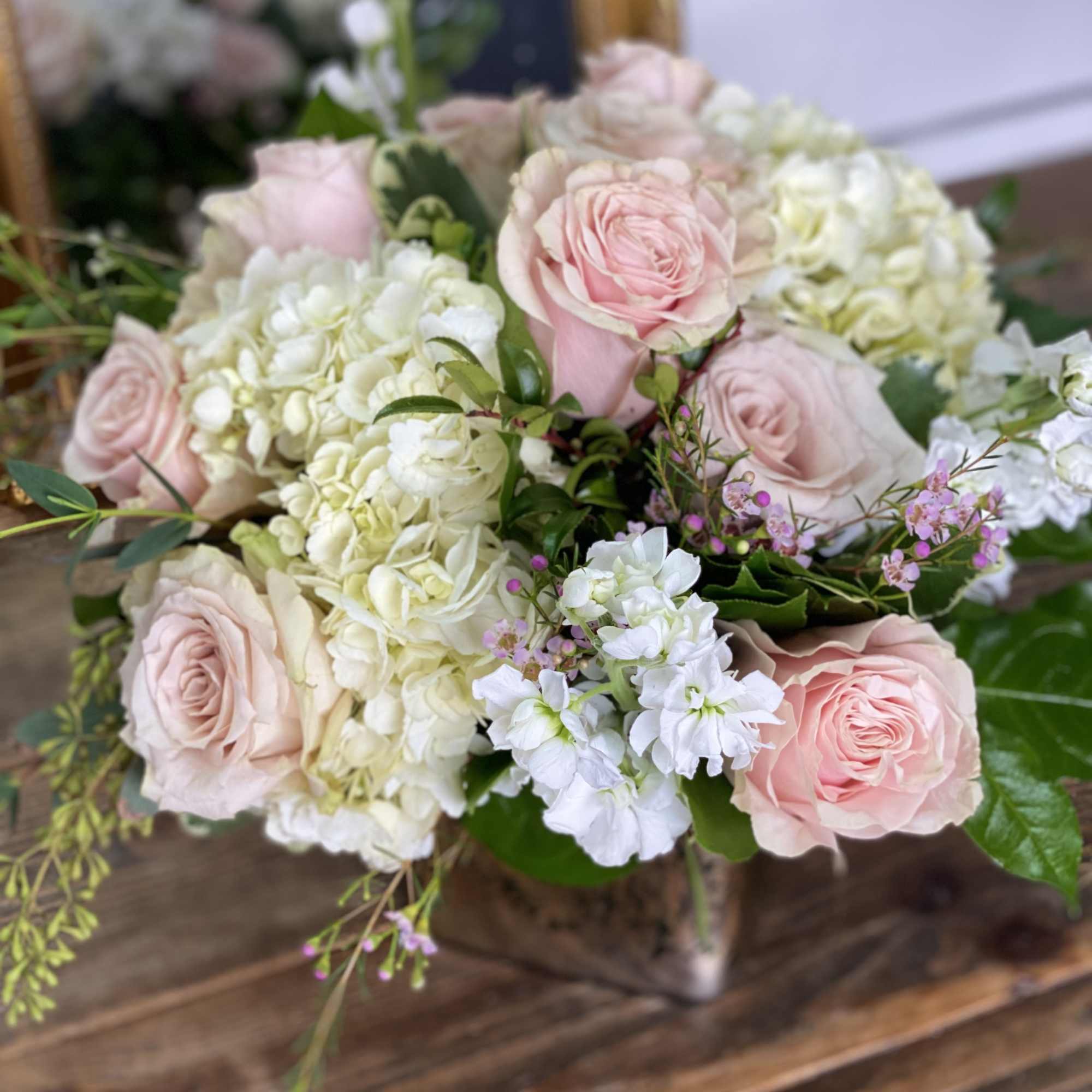 Low arrangement of blush pink roses, white hydrangeas, and white stock in a metallic cube vase on a wooden table.