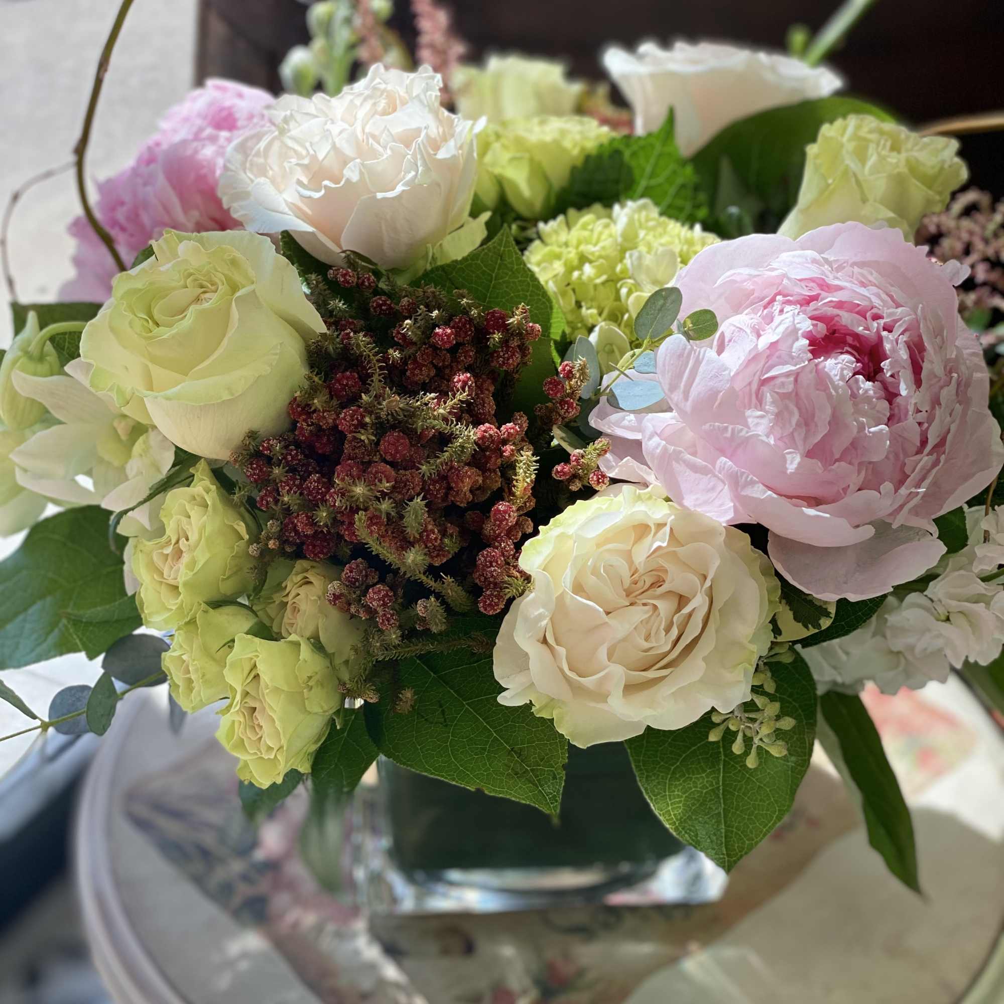 Low arrangement of pale roses, green hydrangeas, and pink peonies in a square glass vase