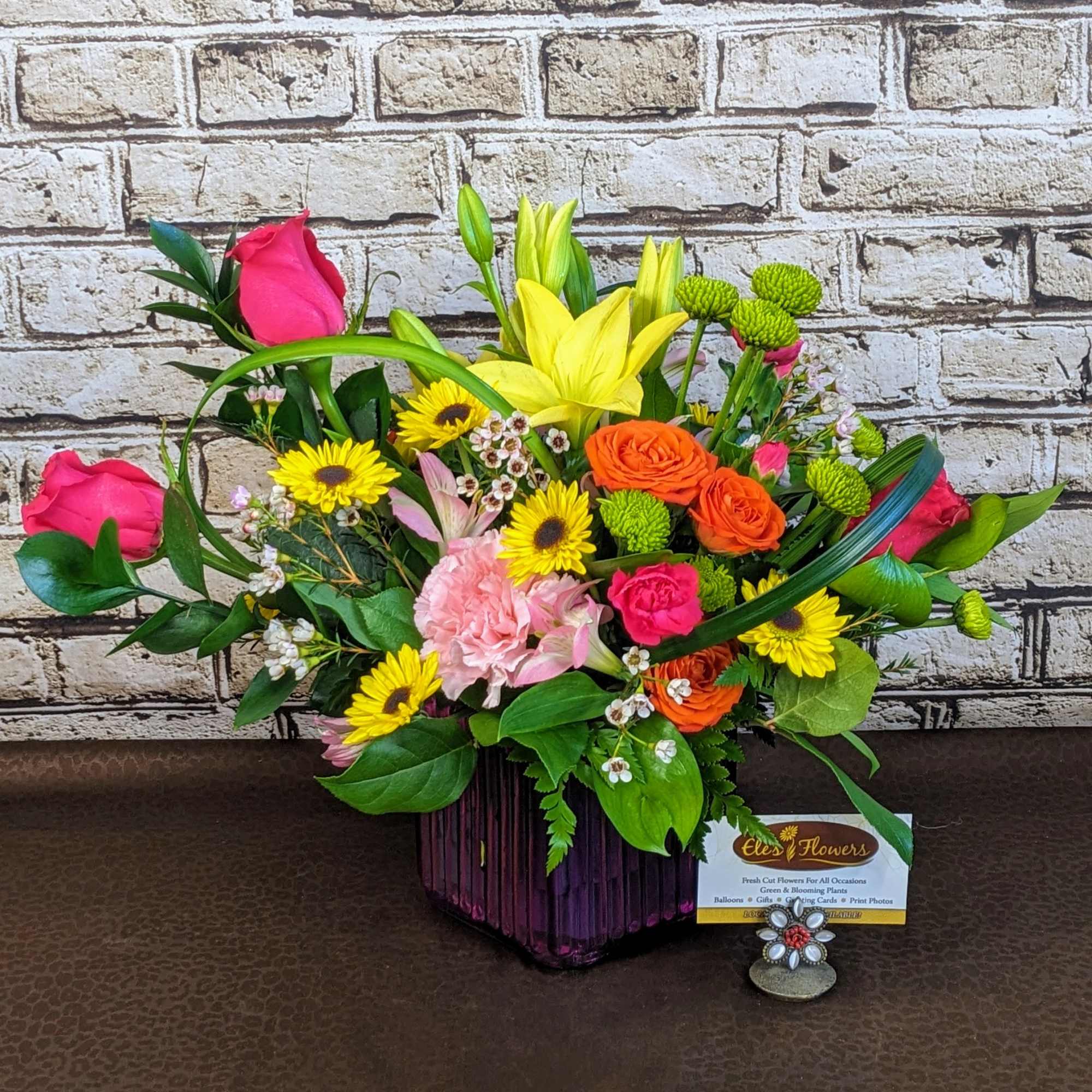 Bright mixed arrangement of yellow lilies and daisies with pink and orange roses in a purple glass vase