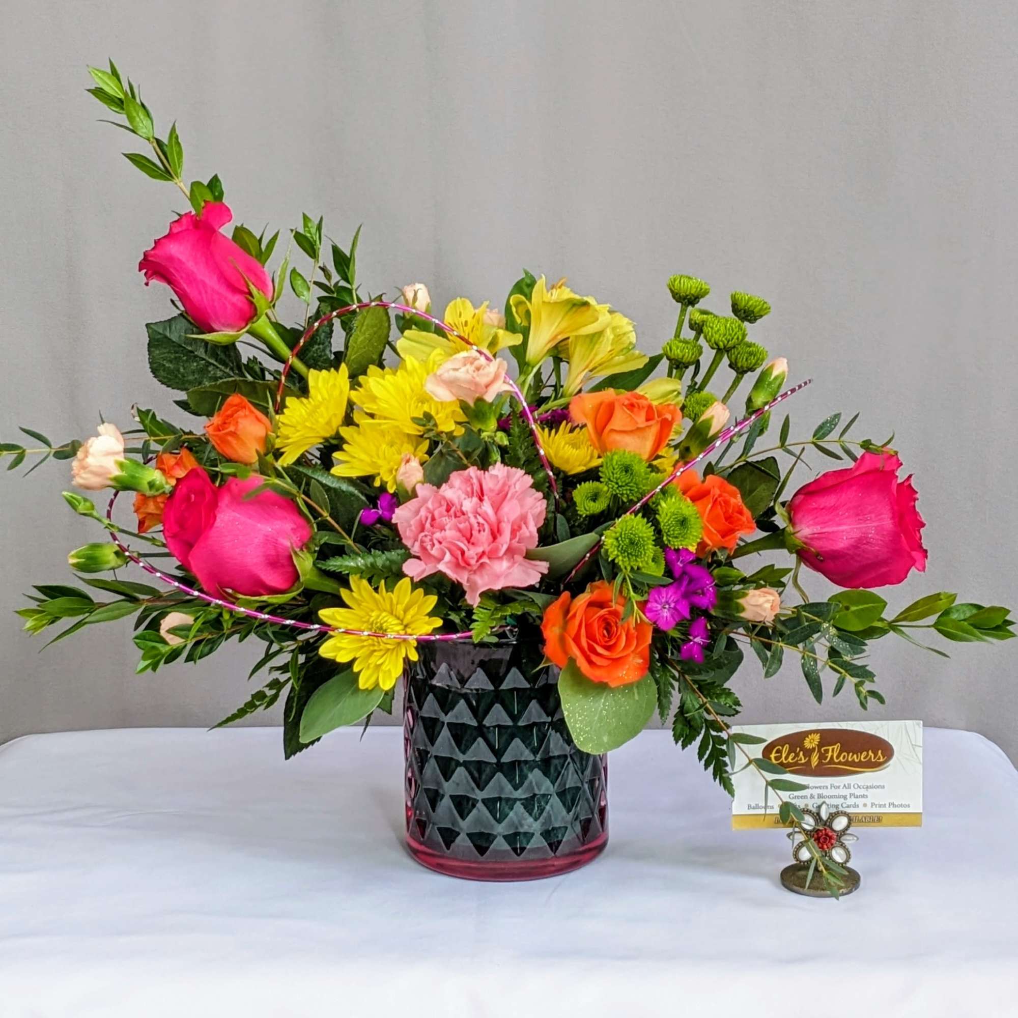 Bright mixed arrangement with pink roses, orange roses, yellow mums, and green button mums in a dark patterned glass vase