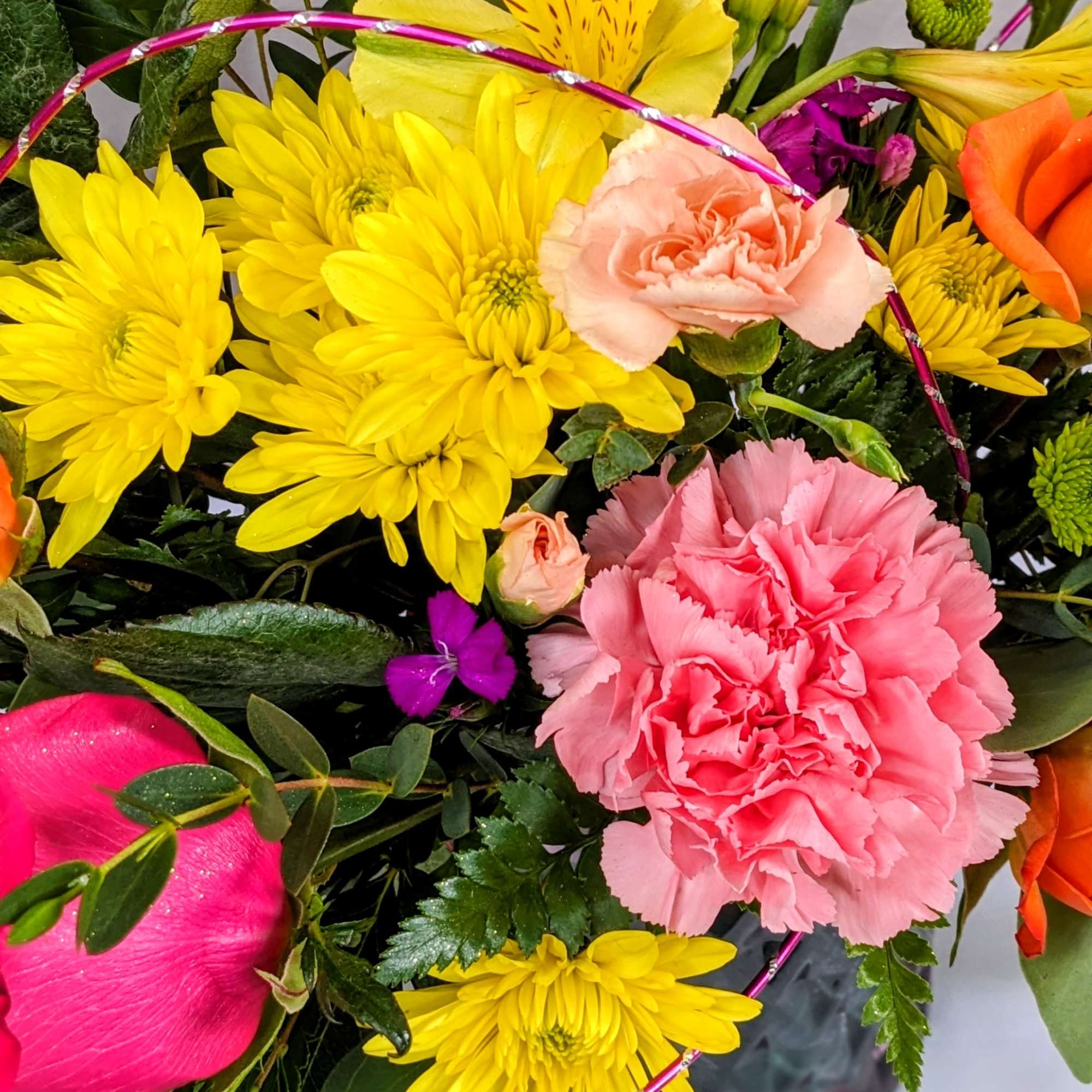 Bright mixed bouquet of yellow mums, pink carnations, and roses in a glass vase with a pink decorative hoop.